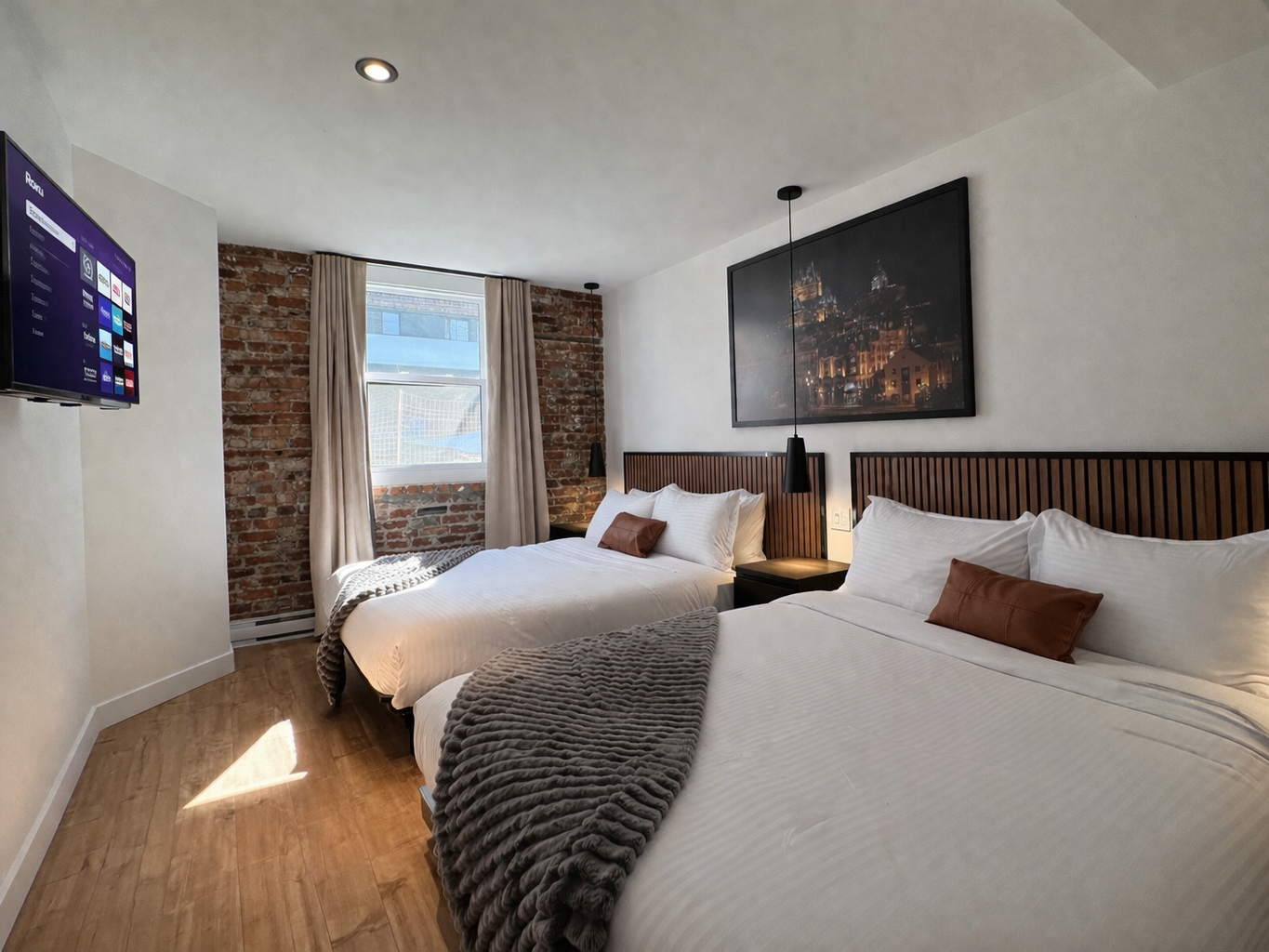 Chambre avec deux lits à linge blanc, tête de lit en bois, tableau de nuit, mur en briques derrière la fenêtre, rideaux beige, télévision à gauche
