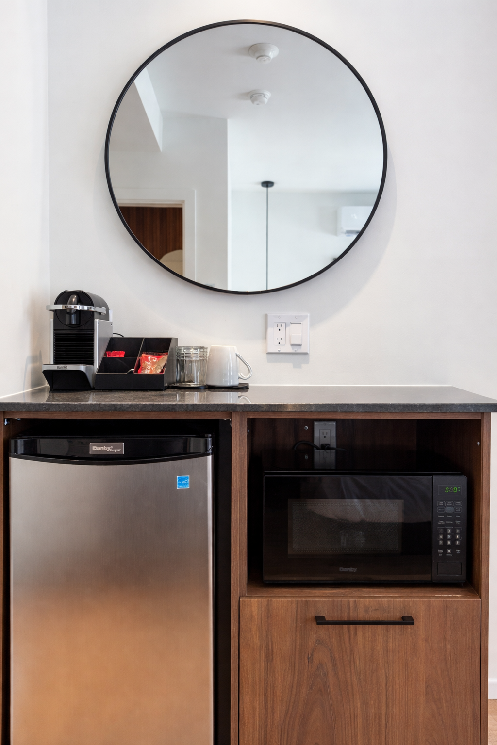 Coin kitchenette avec un miroir rond au mur, un micro-ondes, un mini-réfrigérateur, une machine à café, des tasses, un verre, une prise électrique, et un interrupteur.