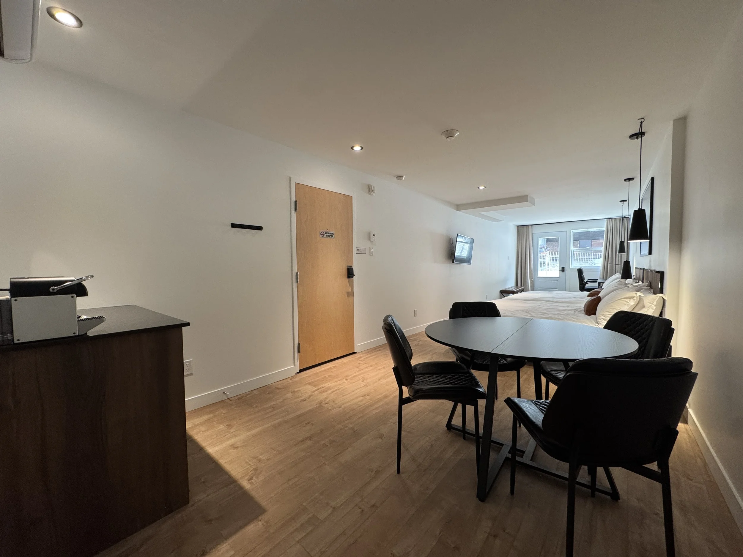 Studio moderne avec lit, fauteuils, table ronde, télévision murale et kitchenette en bois, grandes fenêtres avec rideaux.