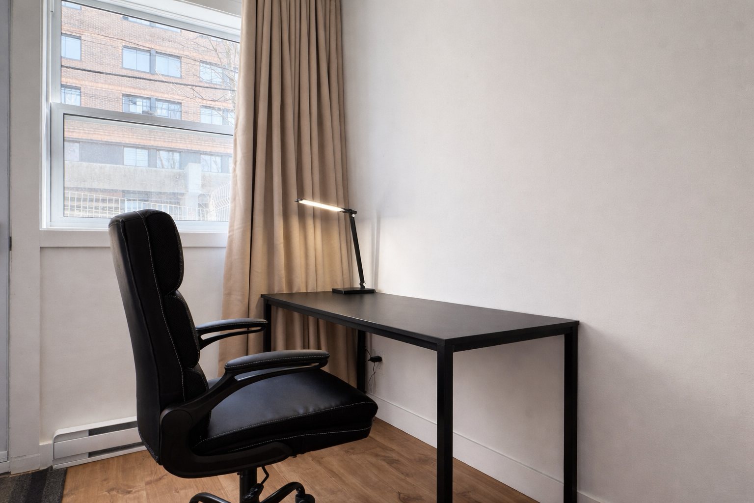 Bureau minimaliste avec une chaise de bureau noire, un bureau en bois noir, une lampe de bureau moderne, une grande fenêtre avec rideaux beige, vue d'un immeuble en brique à l'extérieur.