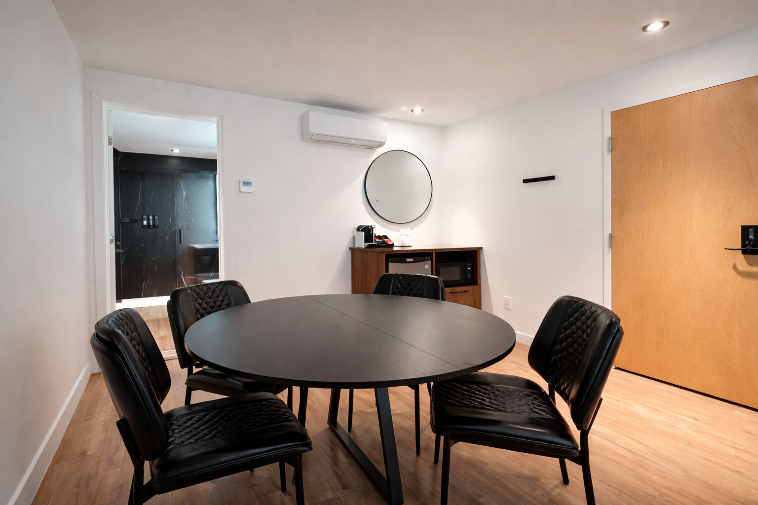 Salle de réunion avec une table ronde noire et six chaises en simili cuir noir, mur blanc avec un grand miroir rond, unité de service en bois avec un micro-ondes et une cafetière, porte en bois clair, porte vitrée derrière apercevant une salle de bai