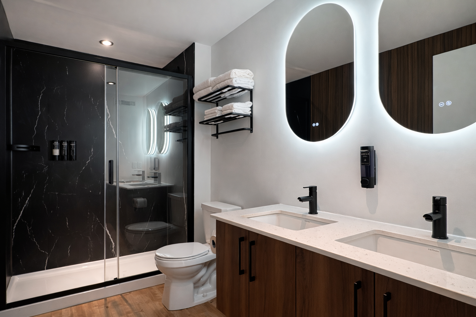Salle de bain avec double lavabo, grands miroirs éclairés, douche vitrée en marbre noir, toilettes, étagère avec serviettes, miroir rond lumineux