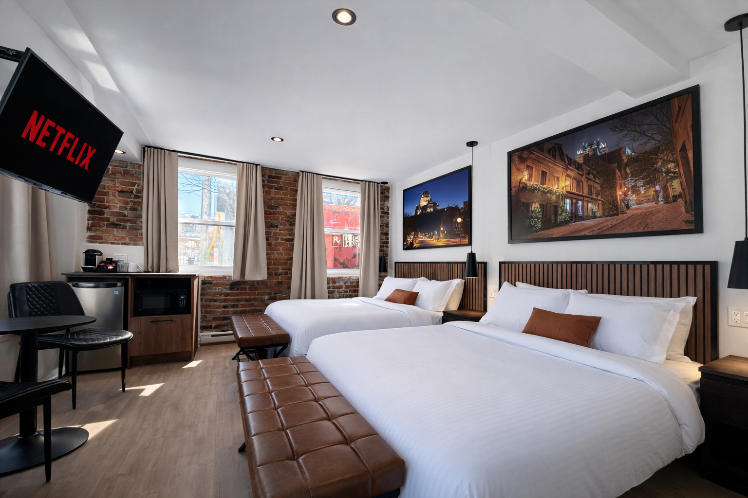 Chambre d'hôtel moderne avec deux lits, œuvres d'art sur le mur, télévision, coin repas avec table et chaises, fenêtres avec rideaux, sol en bois.