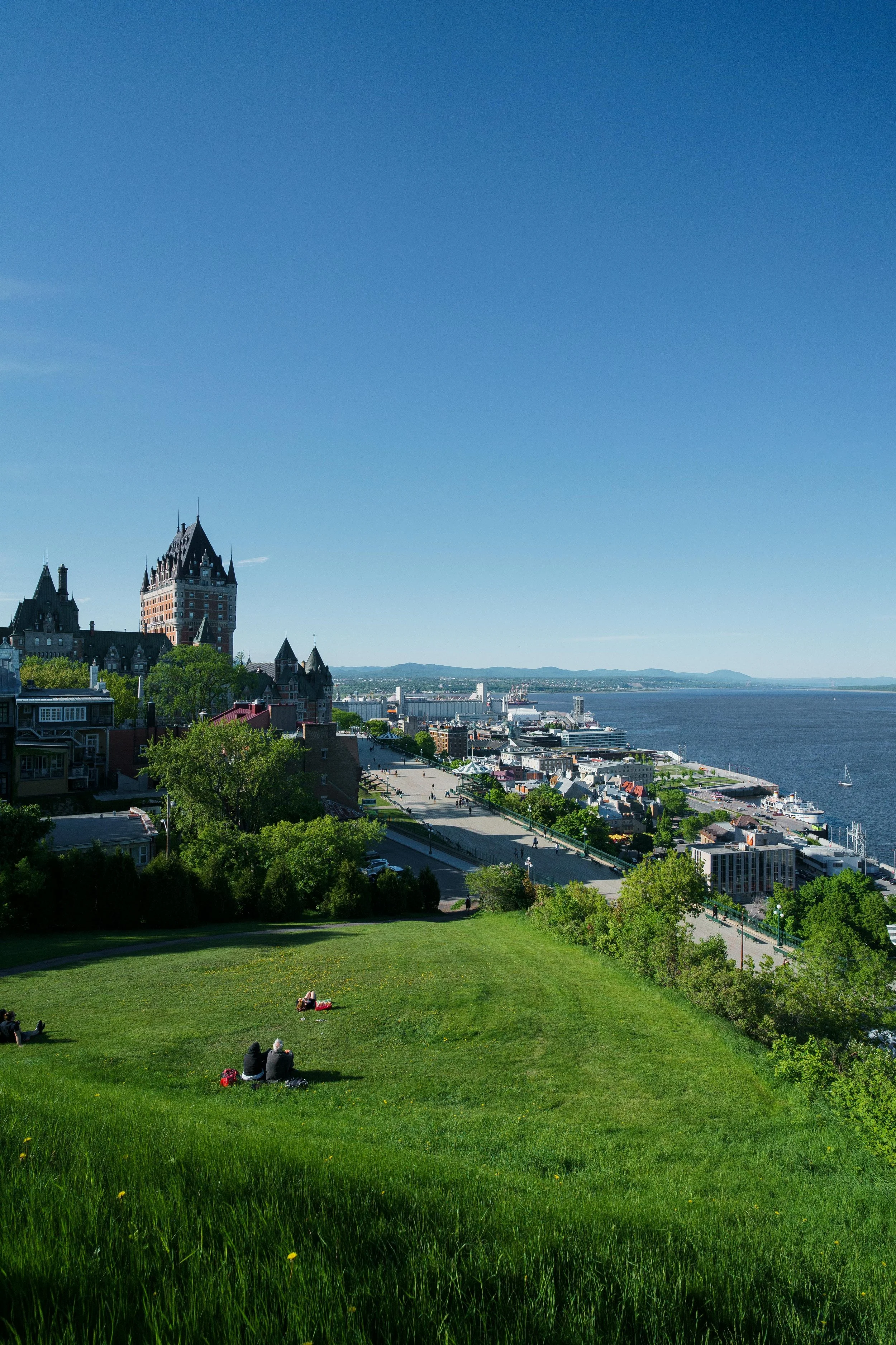 Vue panoramique du Vieux-Québec avec des bâtiments historiques, un parc verdoyant en premier plan, et le fleuve Saint-Laurent en arrière-plan par une journée ensoleillée.