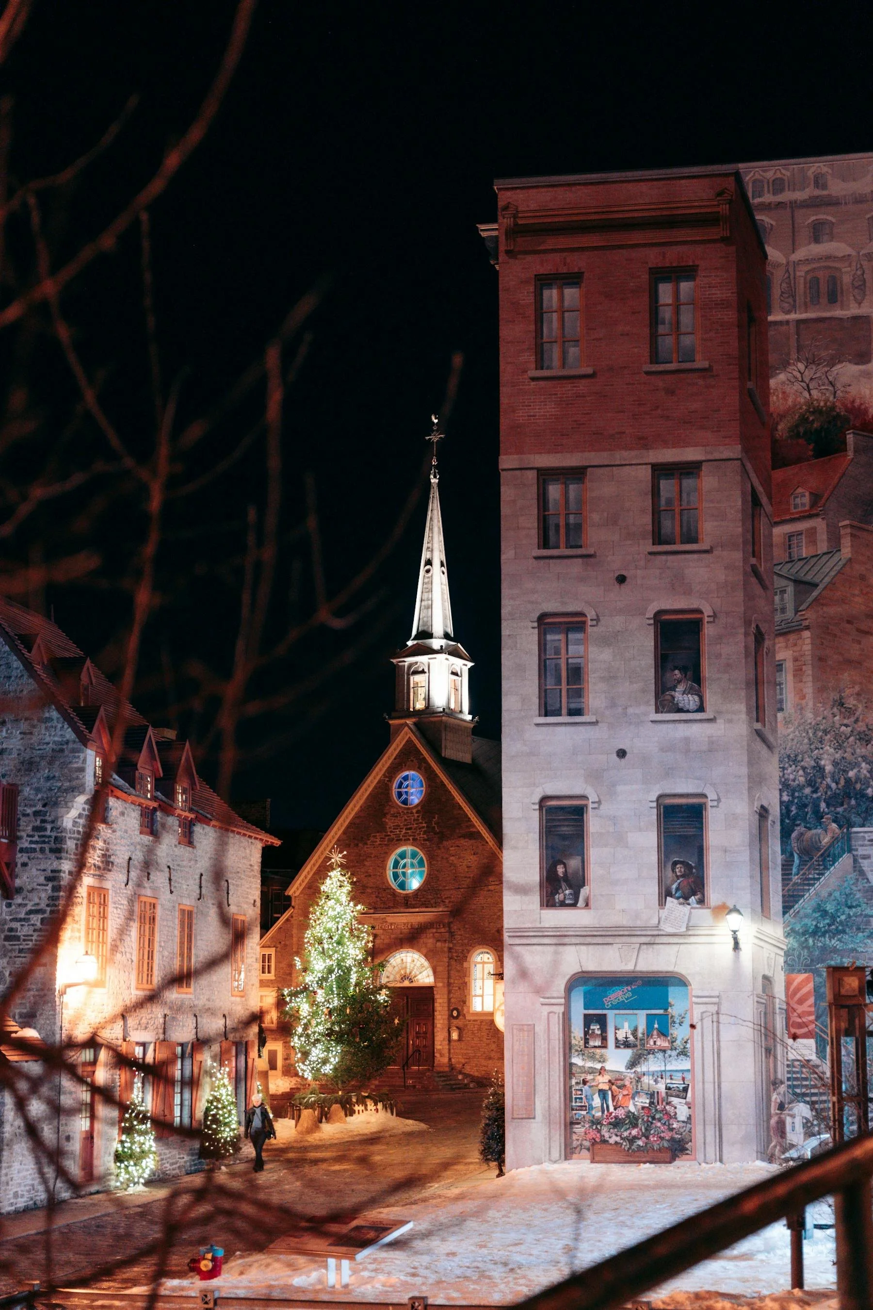 Vue nocturne d'une place de village avec une église illuminée, un arbre de Noël décoré, et des façades de bâtiments ornées de peintures murales. Quelques personnes se promènent dans la rue, ambiance festive de Noël.