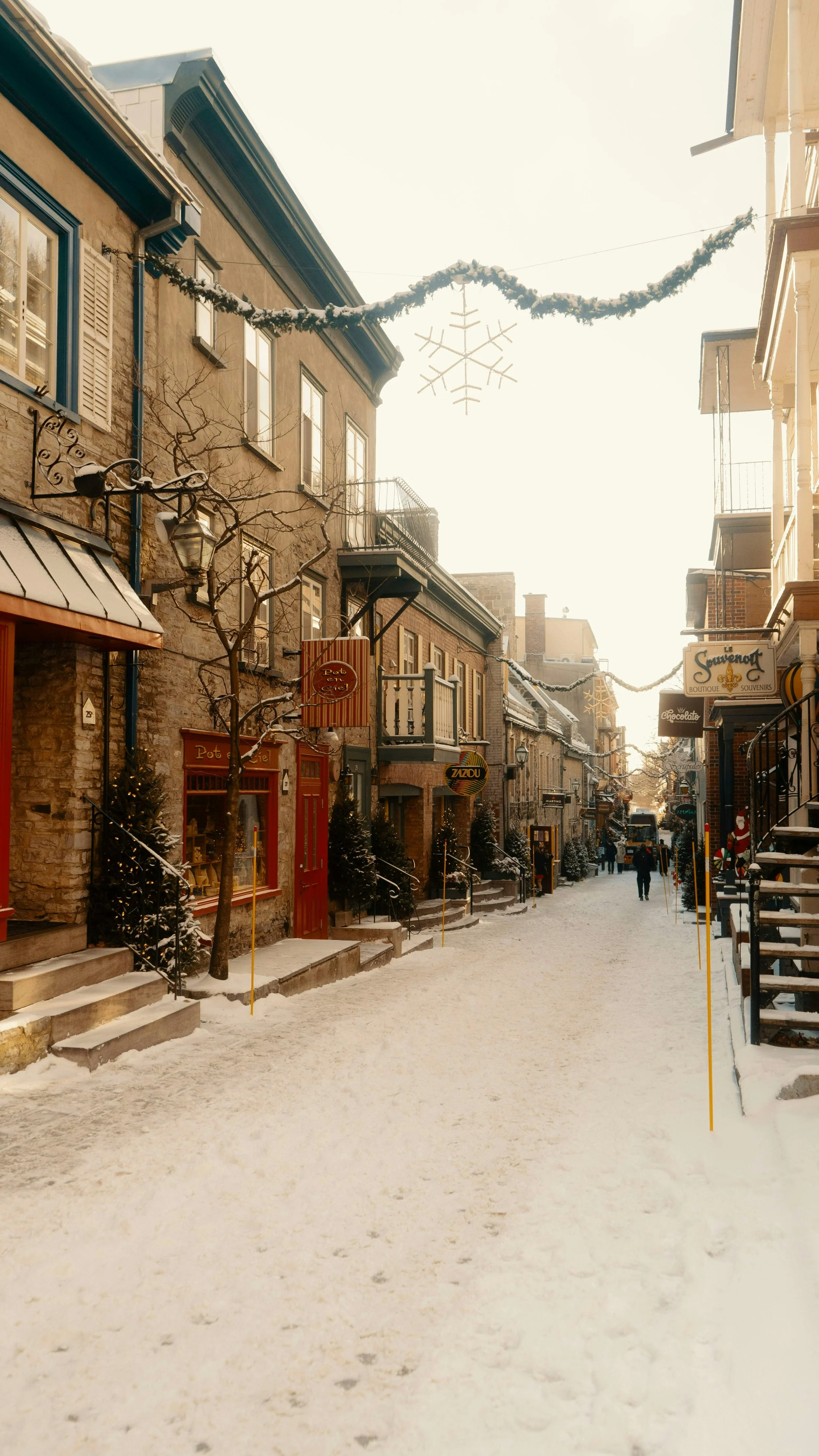 Rue enneigée avec des bâtiments anciens, décorations de Noël suspendues et quelques personnes qui marchent.