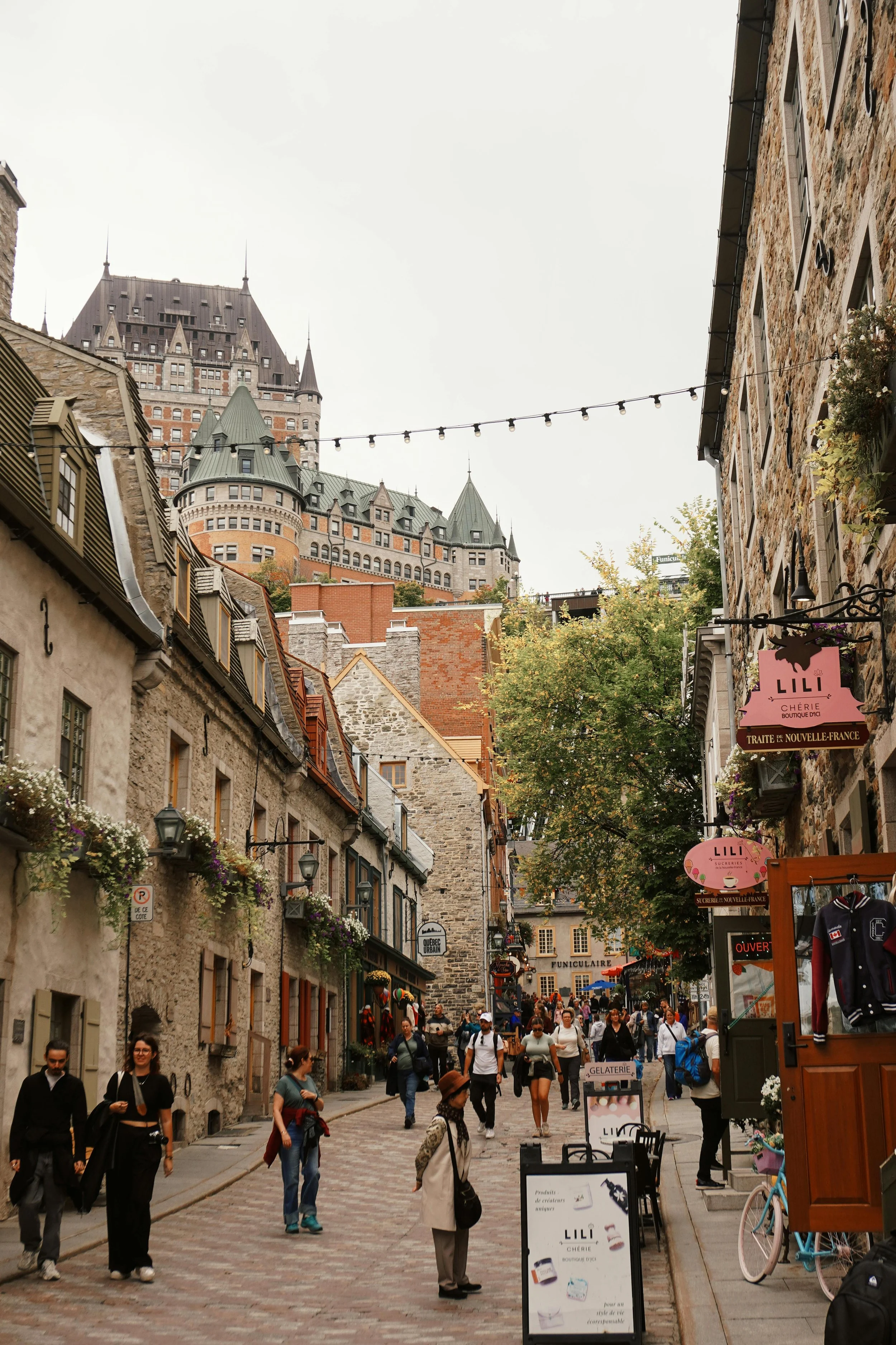 Rue piétonne dans un quartier historique avec des bâtiments en pierre, des magasins et des touristes, avec le Château Frontenac en arrière-plan et des guirlandes lumineuses suspendues au-dessus de la rue.