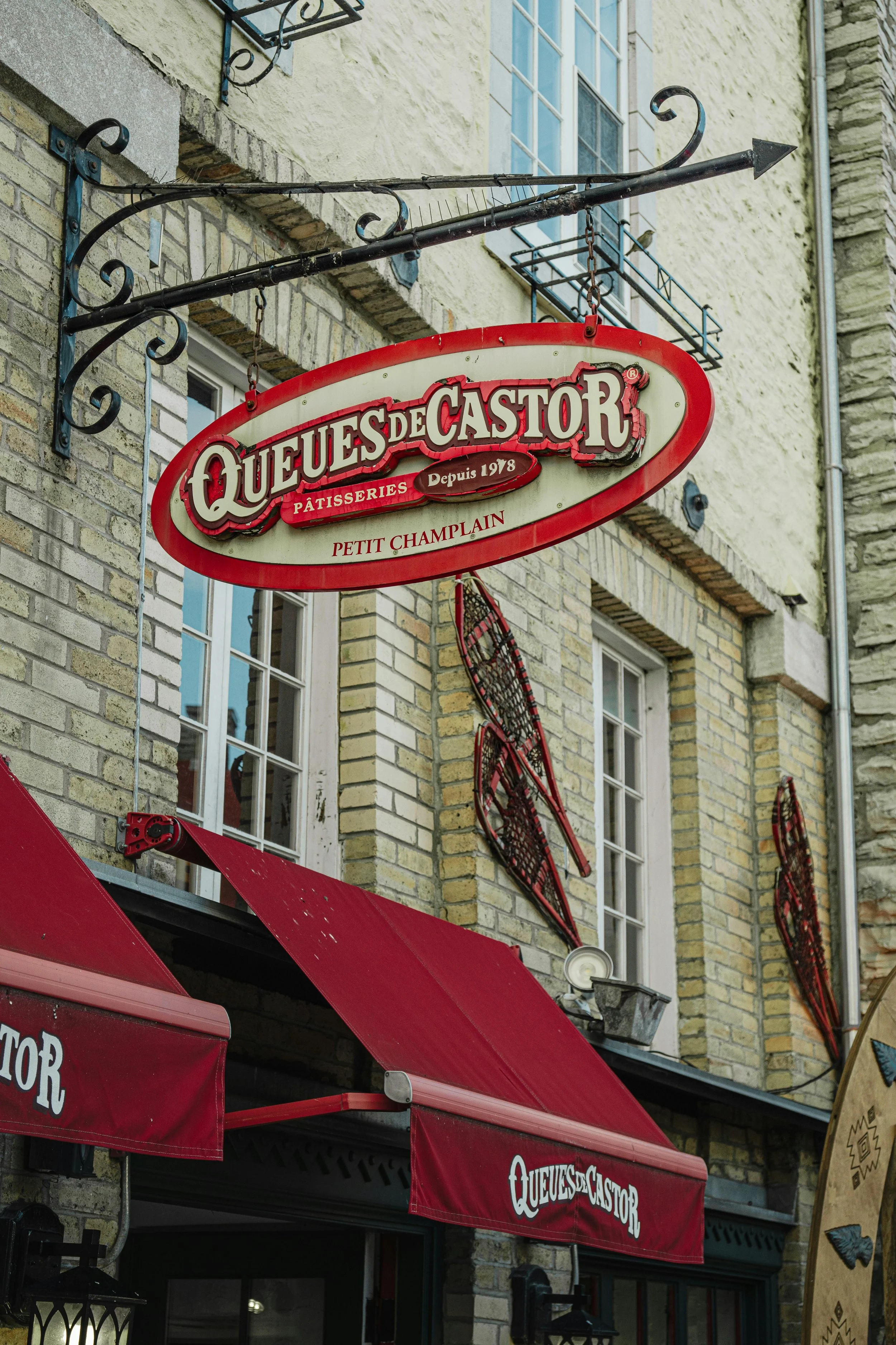 Signalétique de pâtisserie pour 'Quesues de Castor', fondée en 1978, avec une enseigne en forme d'ours en bois, fenêtres avec volets, et décorations de skis en métal sur la façade en pierre.