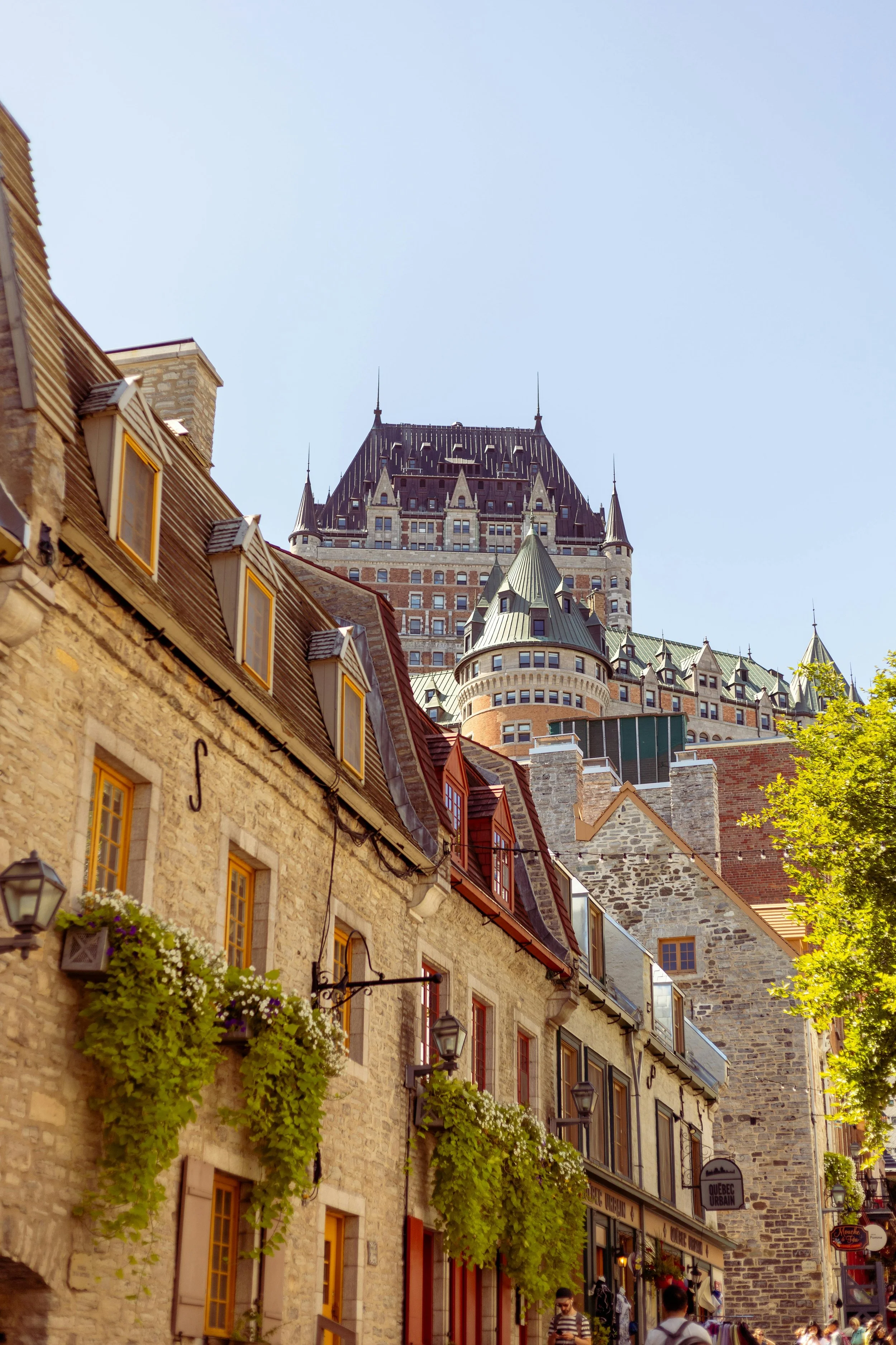 Scène urbaine avec un bâtiment historique Château Frontenac à Québec, avec des maisons anciennes en pierre et des boutiques en premier plan, sous un ciel clair.