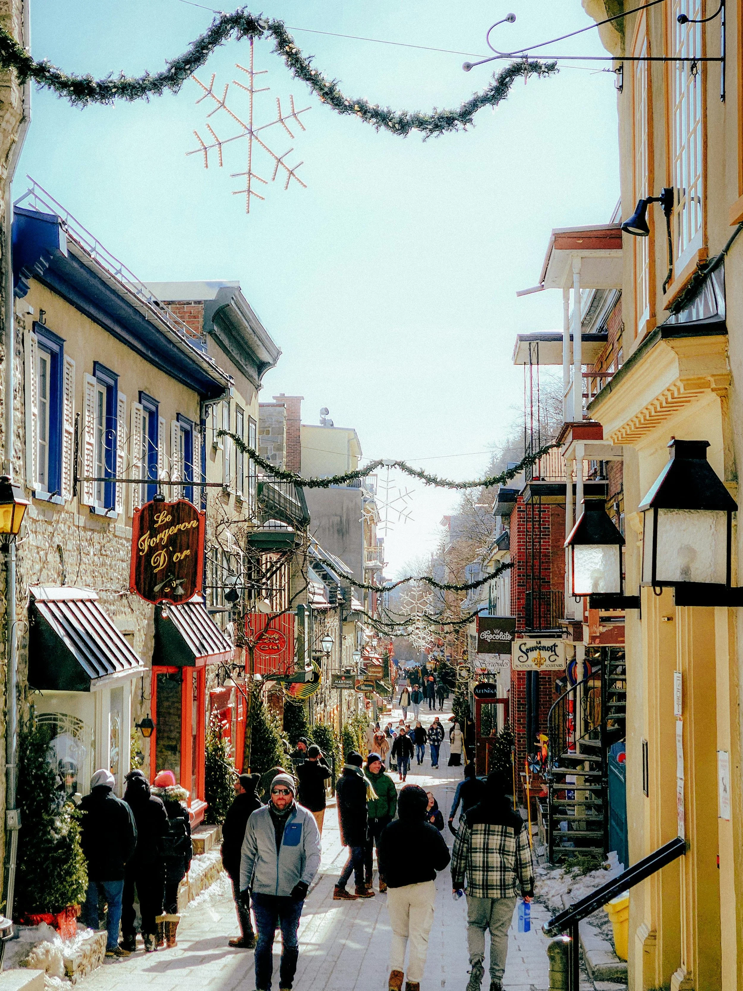 Rue commerçante pavée avec des gens marchant, décorations de Noël suspendues, bâtiments colorés avec boutiques et lumières