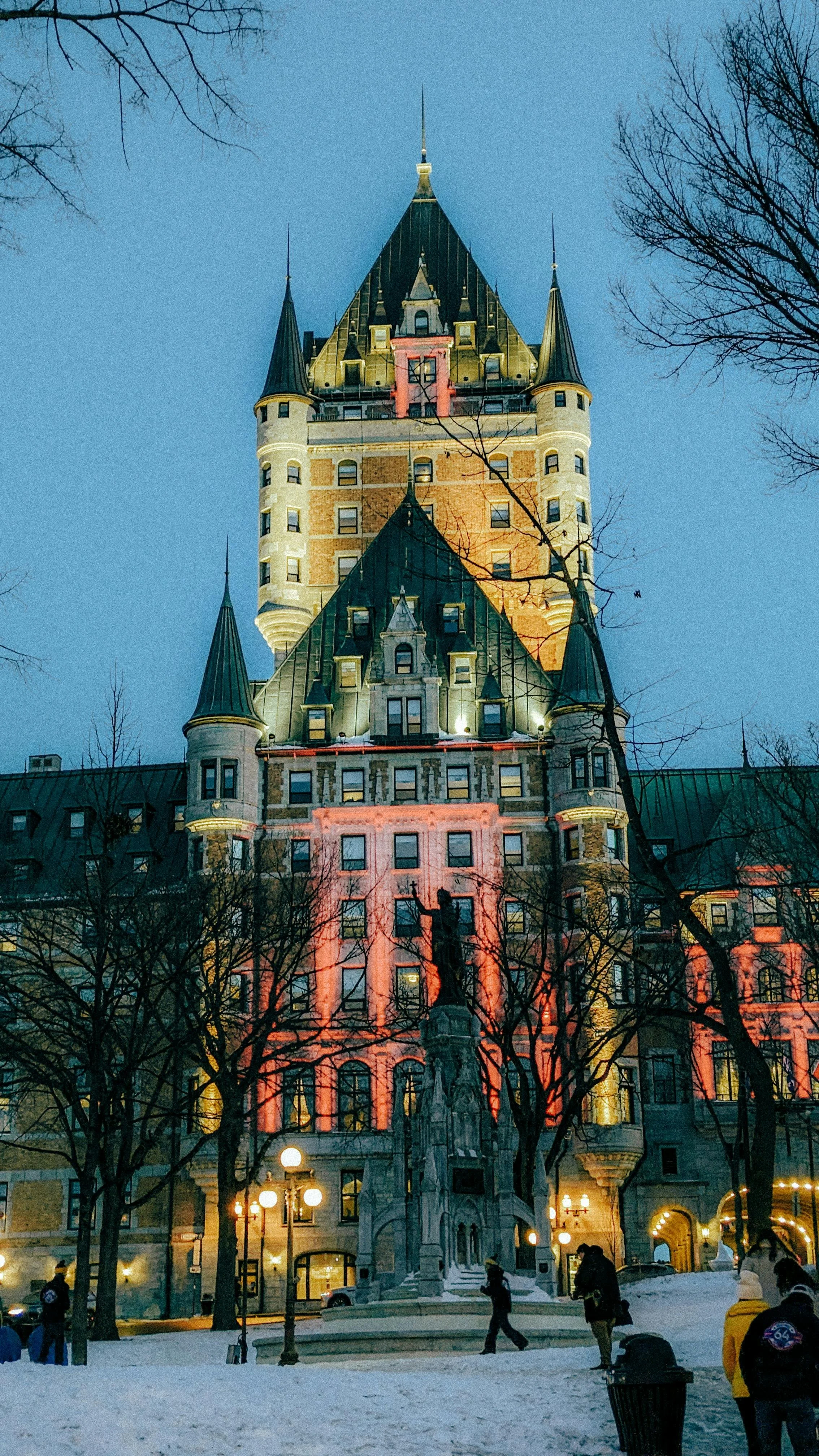 Un bâtiment historique avec des tours pointues, illuminé la nuit, avec des arbres et une place enneigée en premier plan.