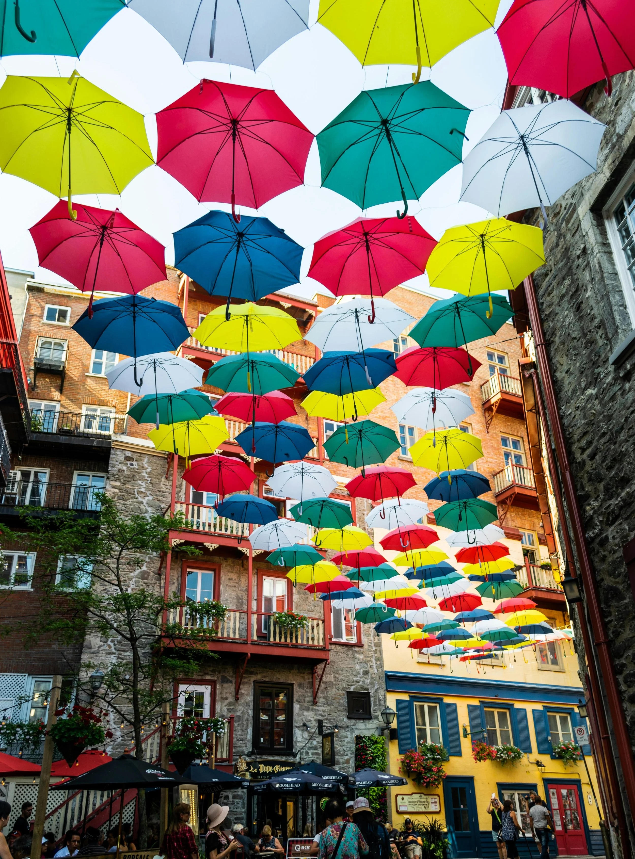 Un ciel rempli de parapluies colorés suspendus au-dessus d'une rue animée, avec des bâtiments en pierre et en brique, et des personnes assises à des cafés.