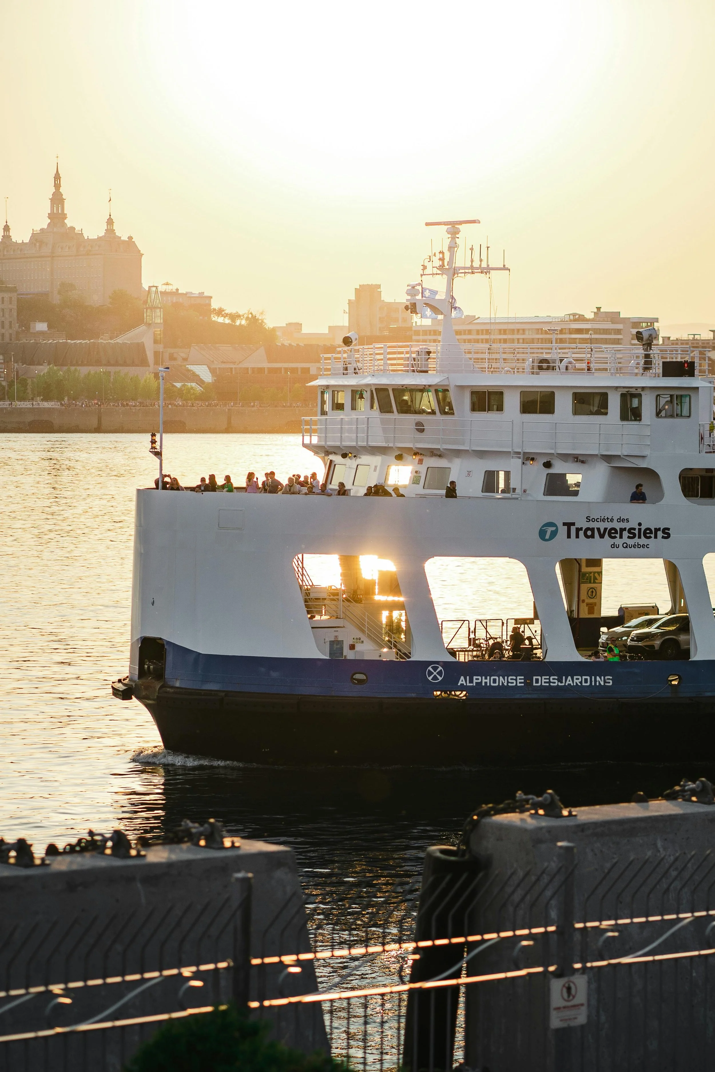 Ferry naviguant sur la rivière au coucher du soleil en ville, avec des passagers à bord et un bâtiment historique en arrière-plan.