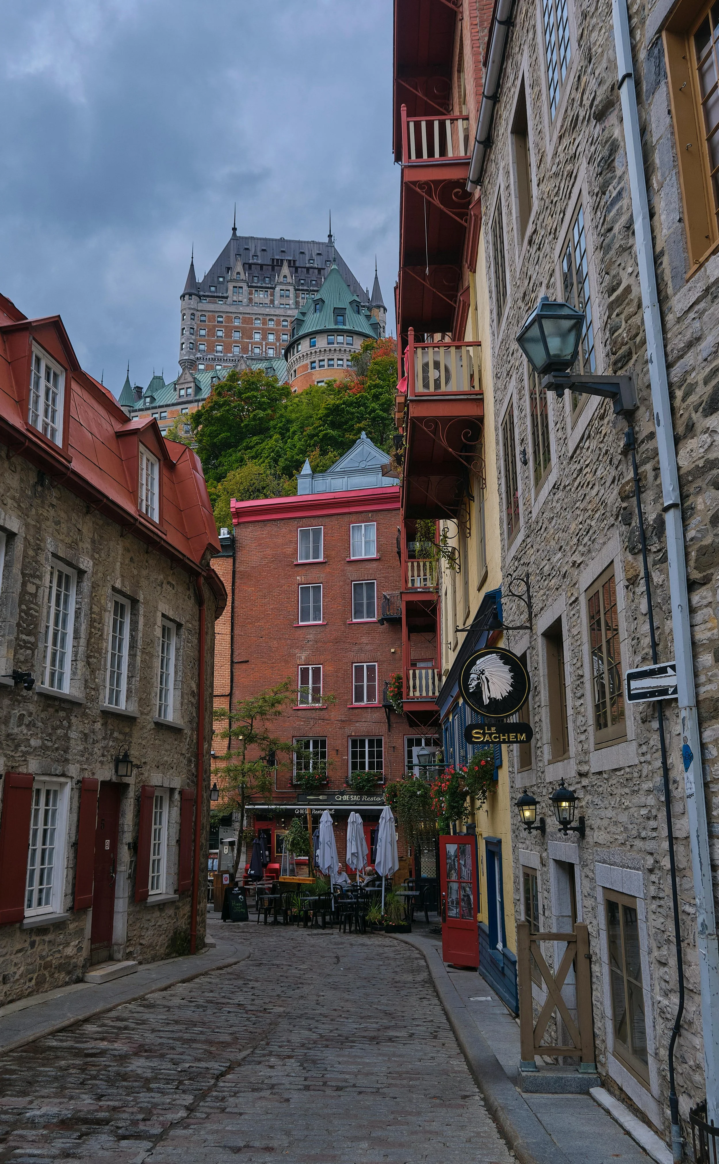 Vue d'une rue pavée bordée de vieux bâtiments en pierre avec des balcons colorés, menant à une terrasse de restaurant avec parasols, et un château en arrière-plan sur une colline.