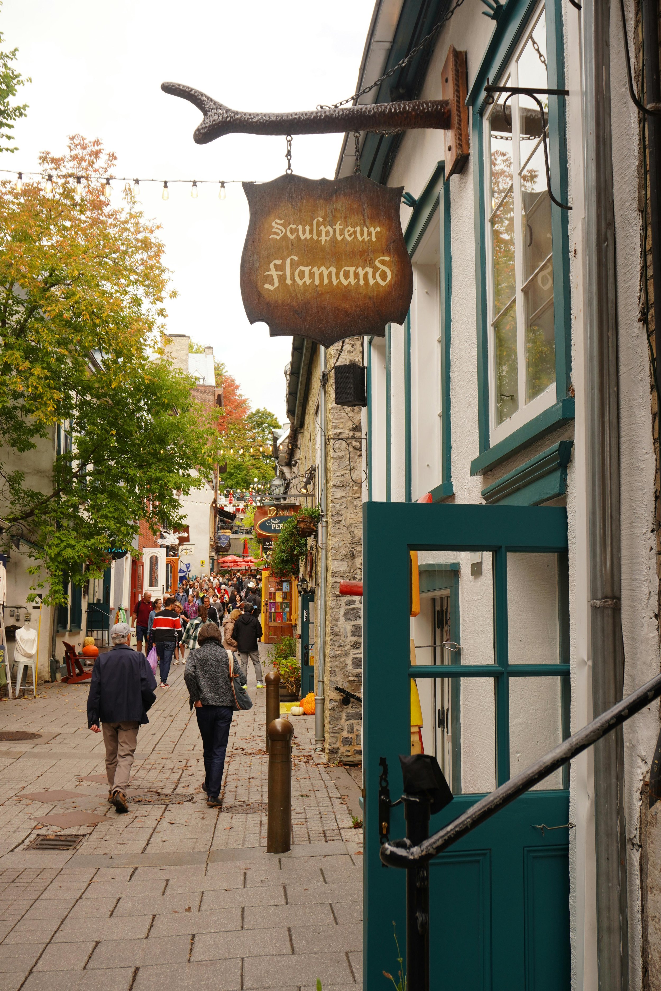 Vue d'une rue piétonne avec des boutiques, des visiteurs et une enseigne en bois indiquant 'Sculpteur flamand' suspendue à une façade de bâtiment dans le centre-ville.