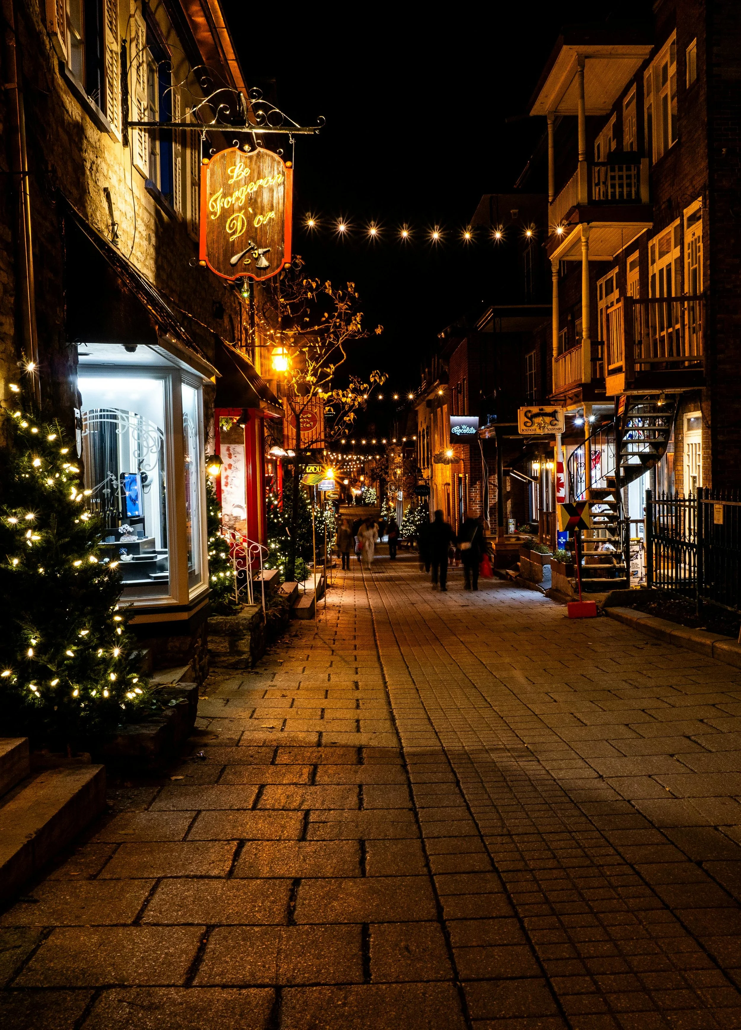 Rue commerçante illuminée la nuit avec des boutiques décorées de lumières de Noël, des passants et un ciel noir.
