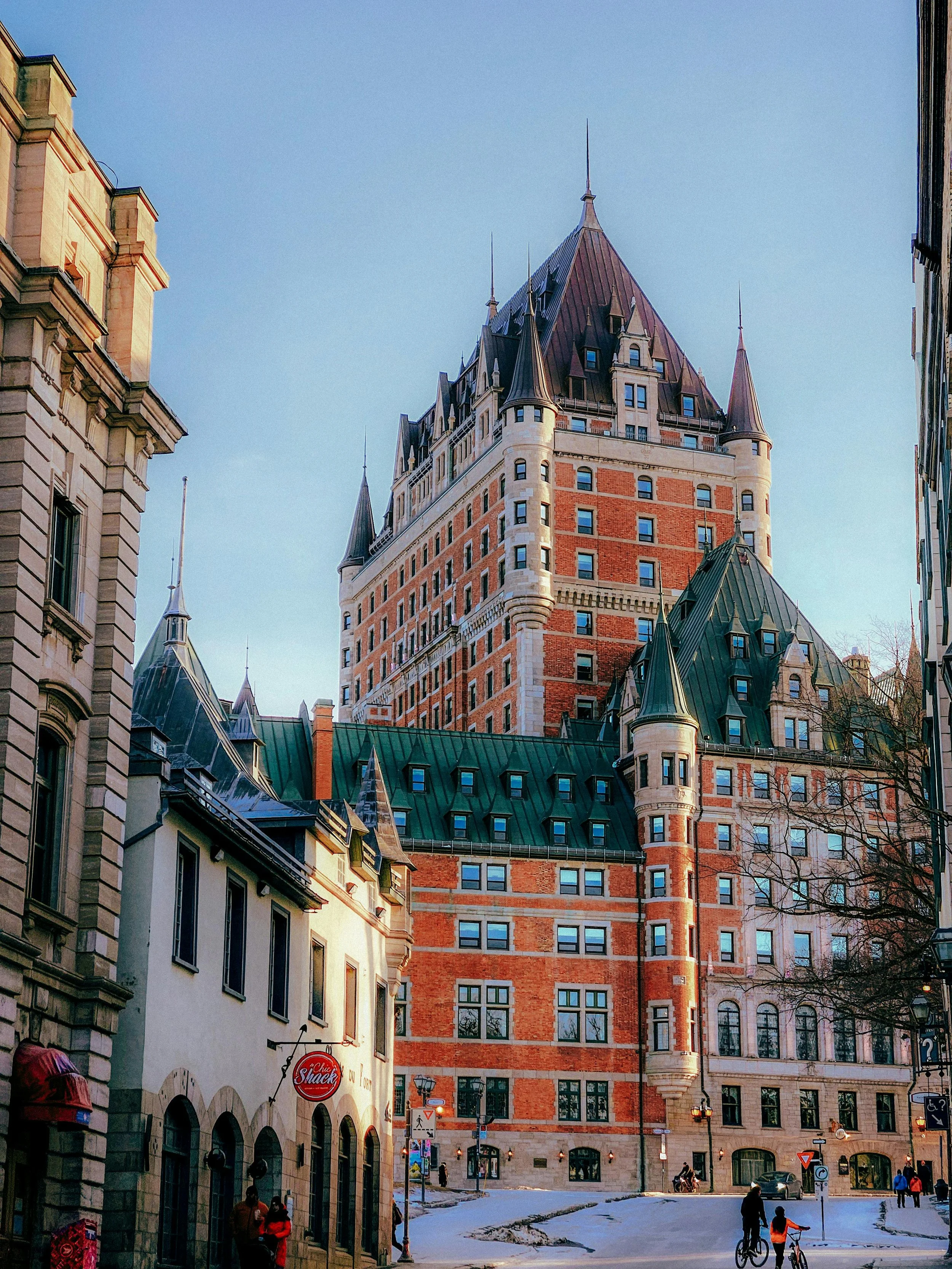 Un immeuble historique en architecture château avec tourelles, murs en brique rouge et ardoise verte, dans une rue animée de Québec avec quelques passants et cyclistes, sous un ciel clair.