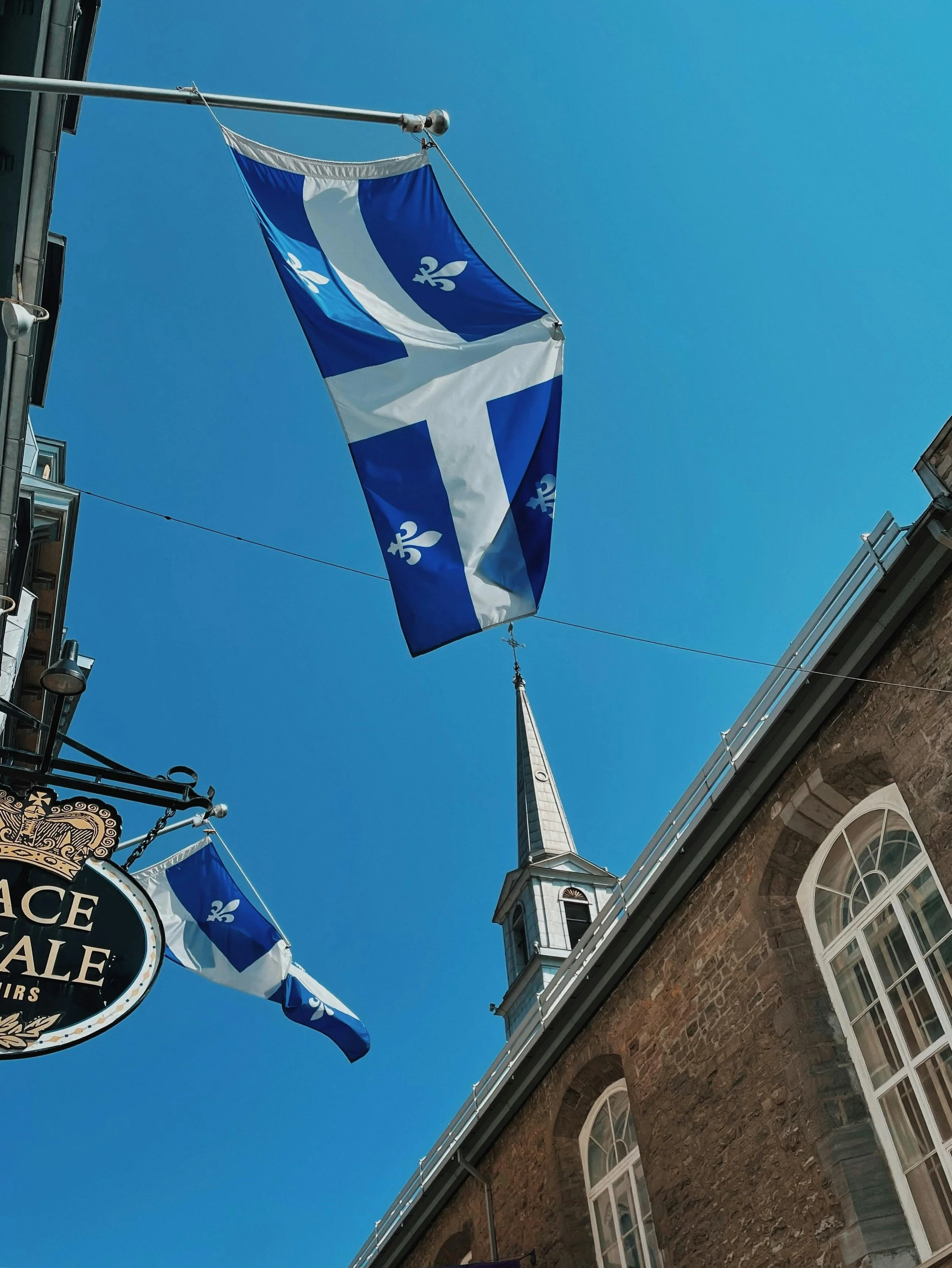 Drapeaux de couleur bleue et blanche avec un symbole en forme de croix ou de fleurs stylisées, flottant dans une rue bordée de bâtiments en pierre, avec une église à clocher en arrière-plan, sous un ciel bleu clair.