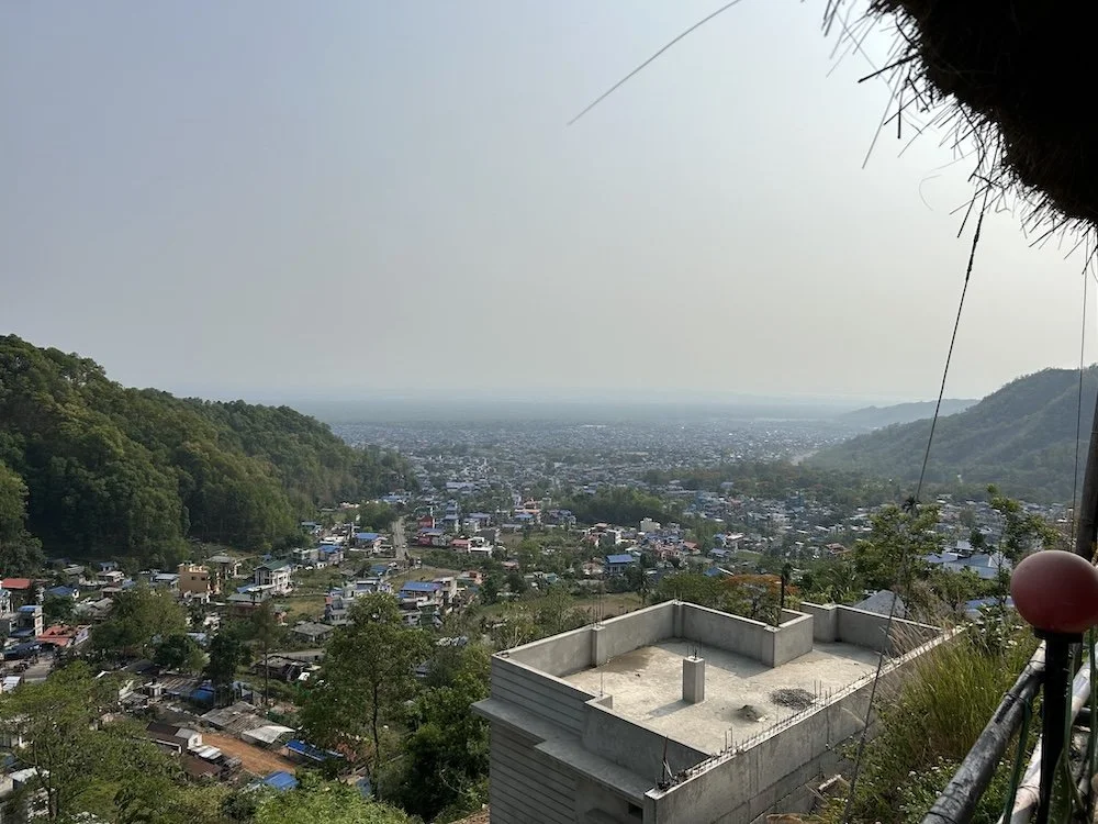 View of a cityscape with densely packed buildings, surrounded by green hills, taken from a hillside vantage point with a partial view of a building rooftop and some wires.
