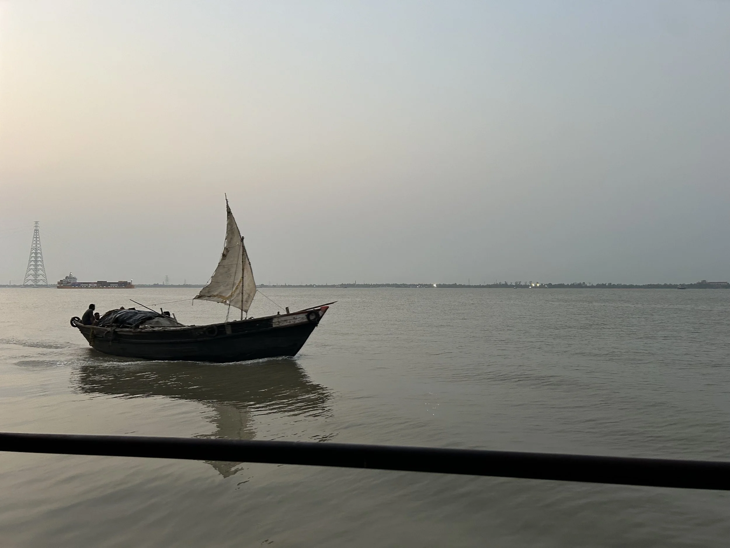 A small wooden boat with a torn sail floating on calm water during daytime with overcast sky, a large cargo ship in the distance, and electrical towers on the horizon.