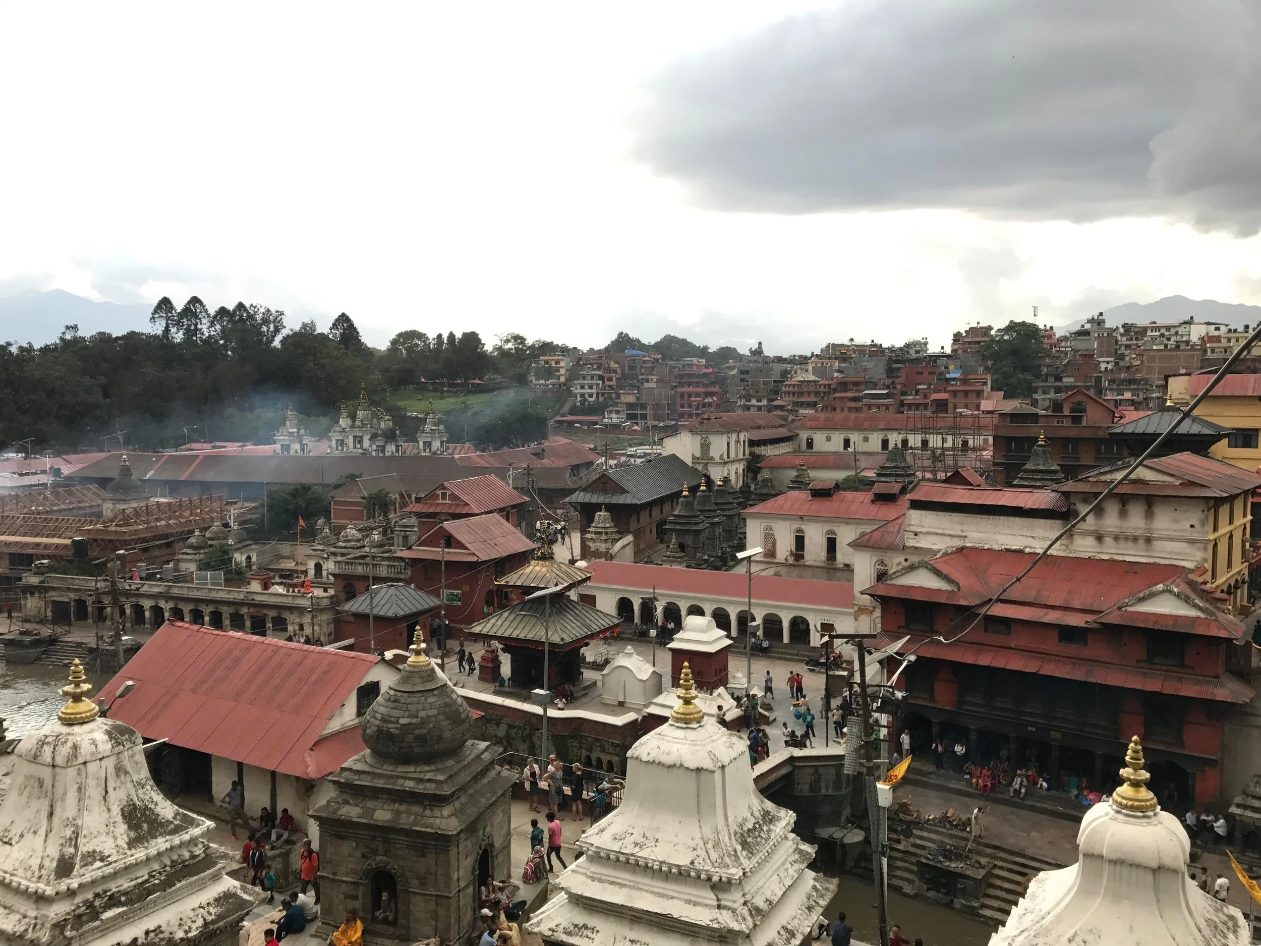 A cityscape of temples and buildings with red roofs in a hilly area, with people walking and traditional structures in the foreground, cloudy sky overhead.