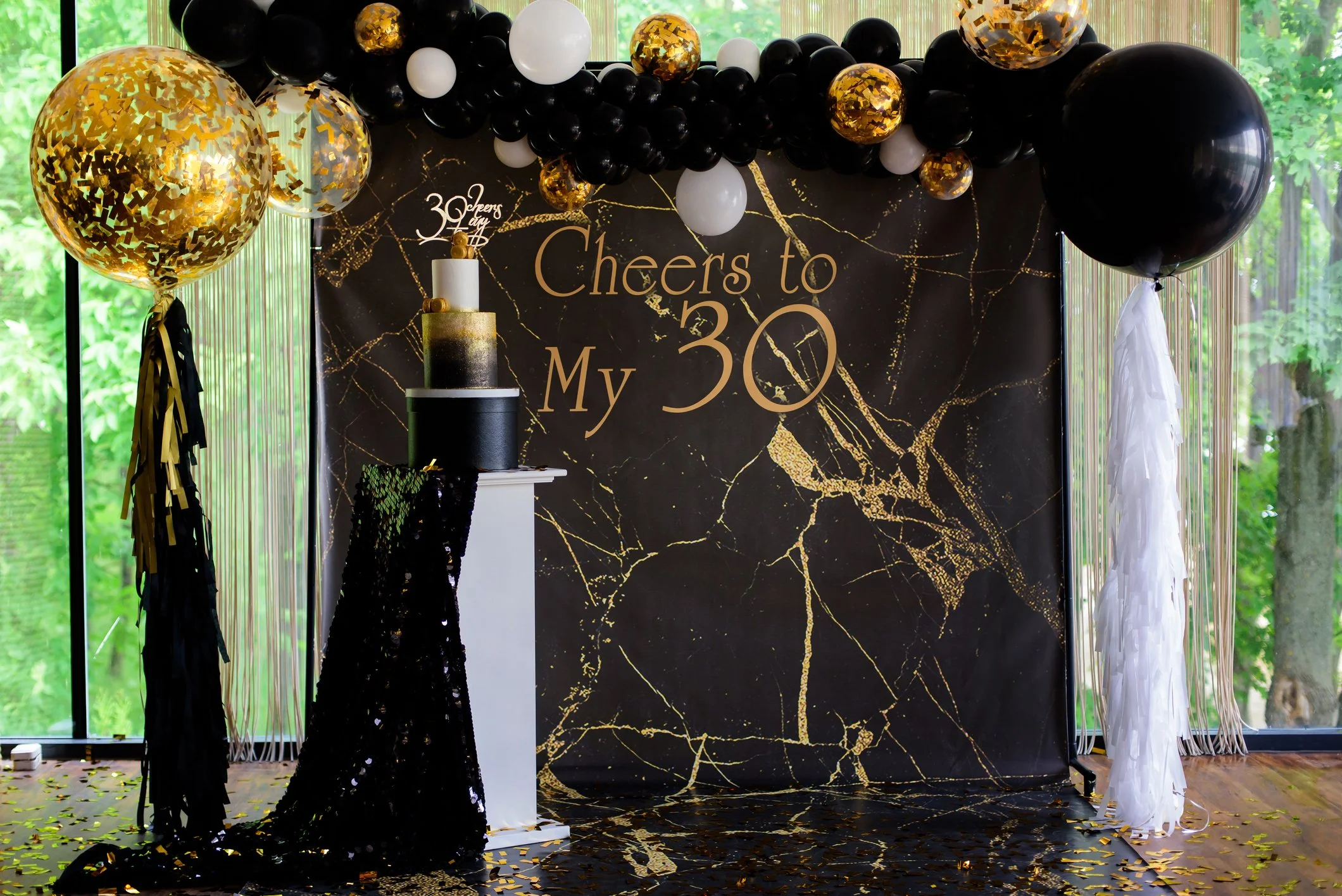 A celebration setup with black, white, and gold balloons arranged in an arch above a black backdrop with gold marble patterns. The backdrop features the text "Cheers to My 30" in gold lettering. There is a tall black and white layered cake with a gold topper that says "30 Cheers." Standing on the floor are two small white pedestals, one draped with a black sequin cloth, and the other with white hanging decorations. Gold confetti is scattered on the wooden floor.
