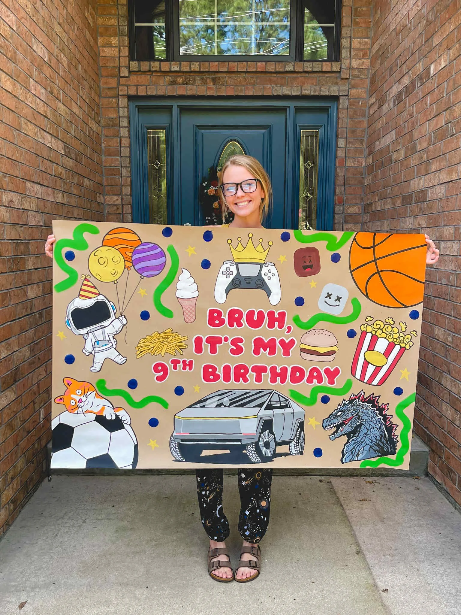 A woman holding a colorful birthday sign outside a house with brick walls and a blue door. The sign features drawings of balloons, a robot game controller, a basketball, popcorn, a hamburger, a soccer ball with a cat, a monster, ice cream, and various decorations. The sign reads 'Bruh, it's my 9th birthday' in large pink letters.