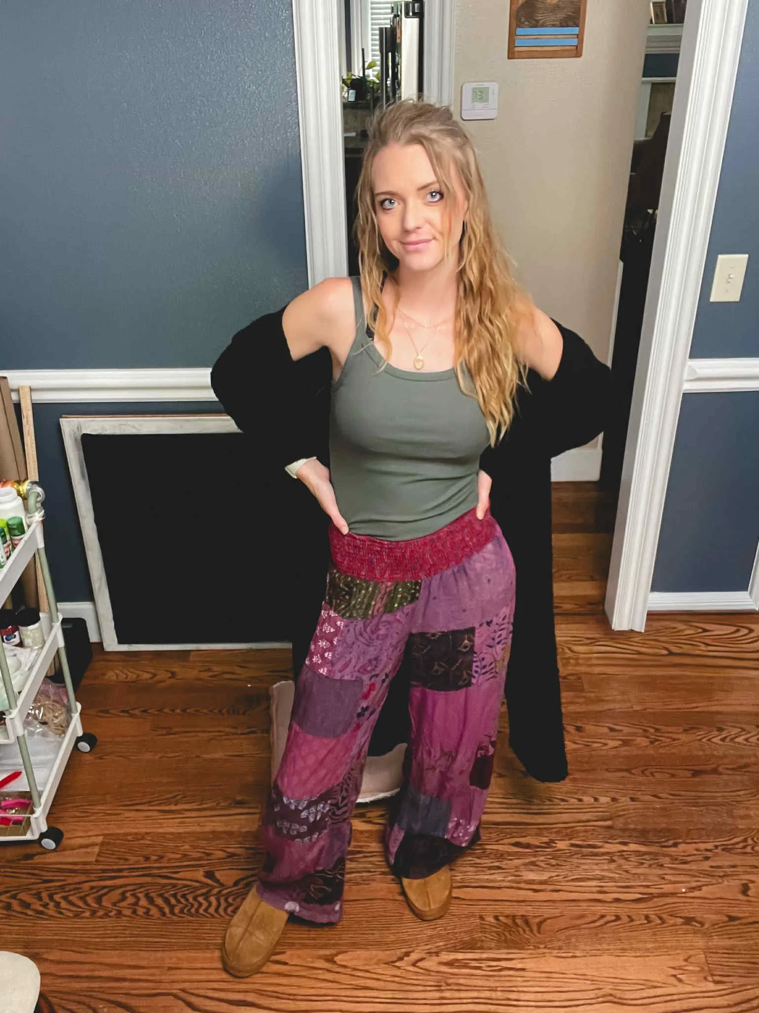 A woman standing indoors on a wooden floor, wearing beige shoes, wide-leg purple patchwork pants, a green tank top, and a black cardigan. She has long, wavy blonde hair and is posing with her hands on her hips, smiling slightly.