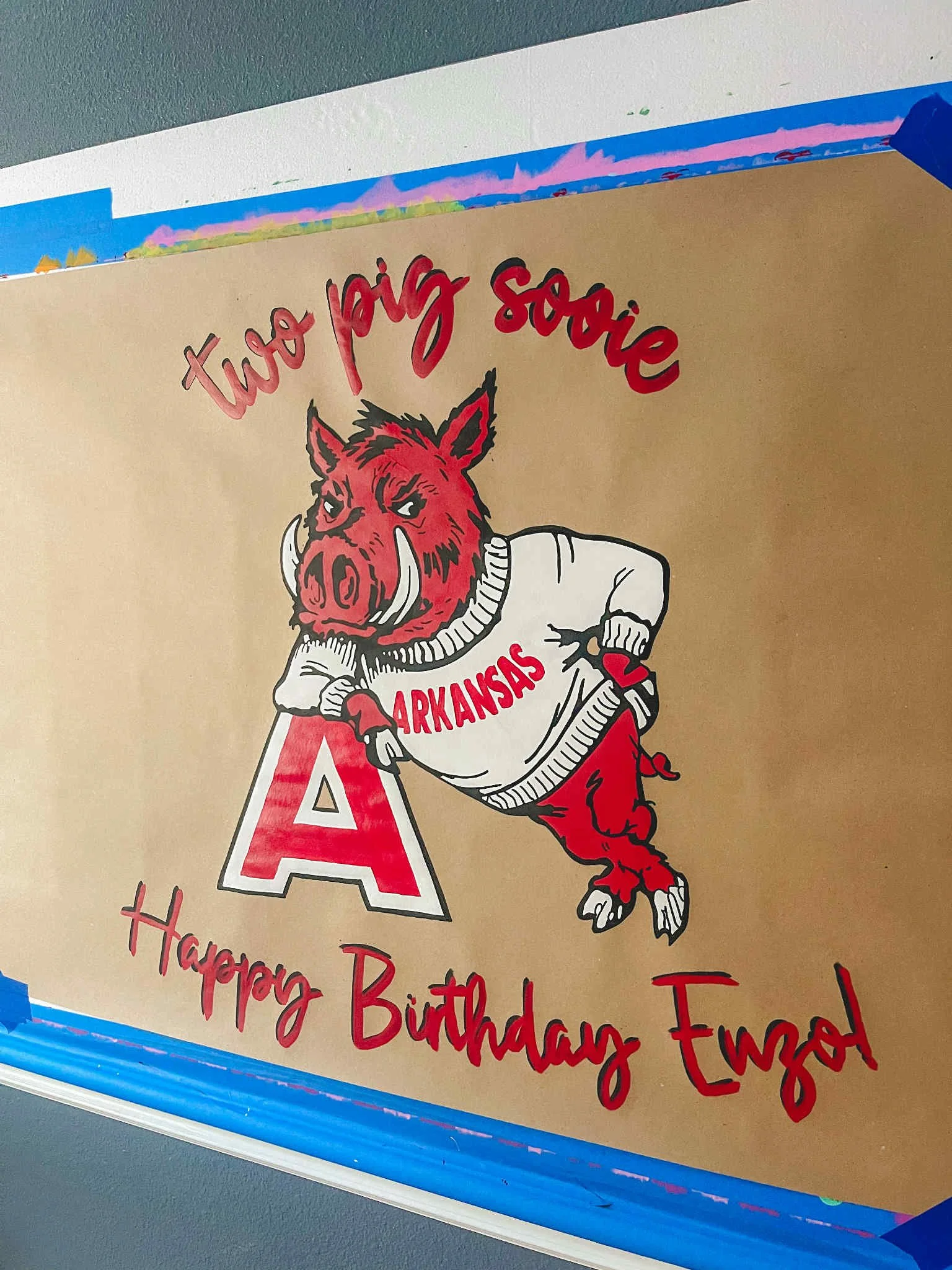 A birthday greeting card with a cartoon boar wearing a white sweater with 'Arkansas' written on it, leaning on a large red letter 'A'. The card has red and black text that says, 'two pig sorie' at the top and 'Happy Birthday Enzo!' at the bottom.