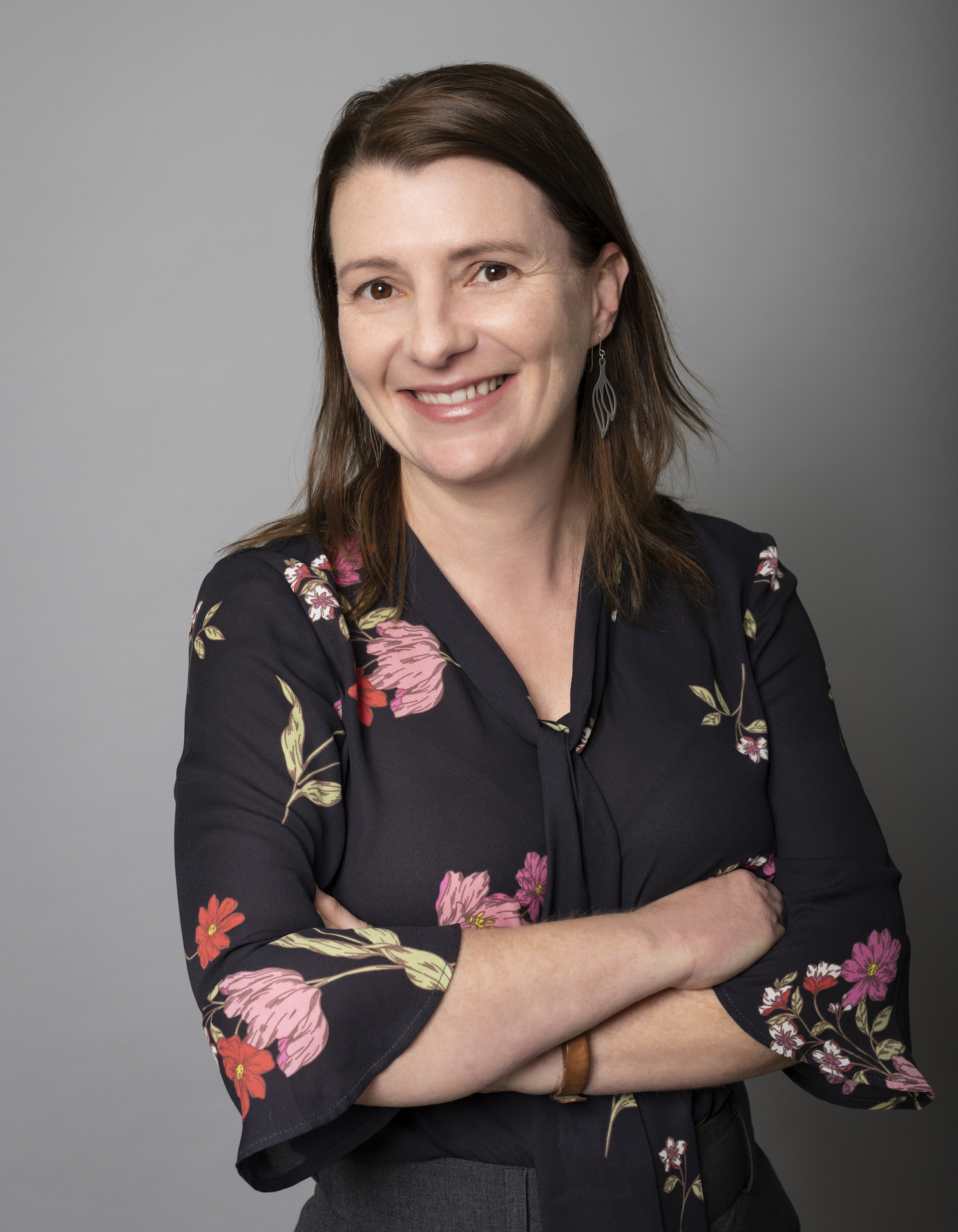 A woman with long brown hair wearing a black floral blouse with pink and red flowers, and earrings, posed against a gray background.