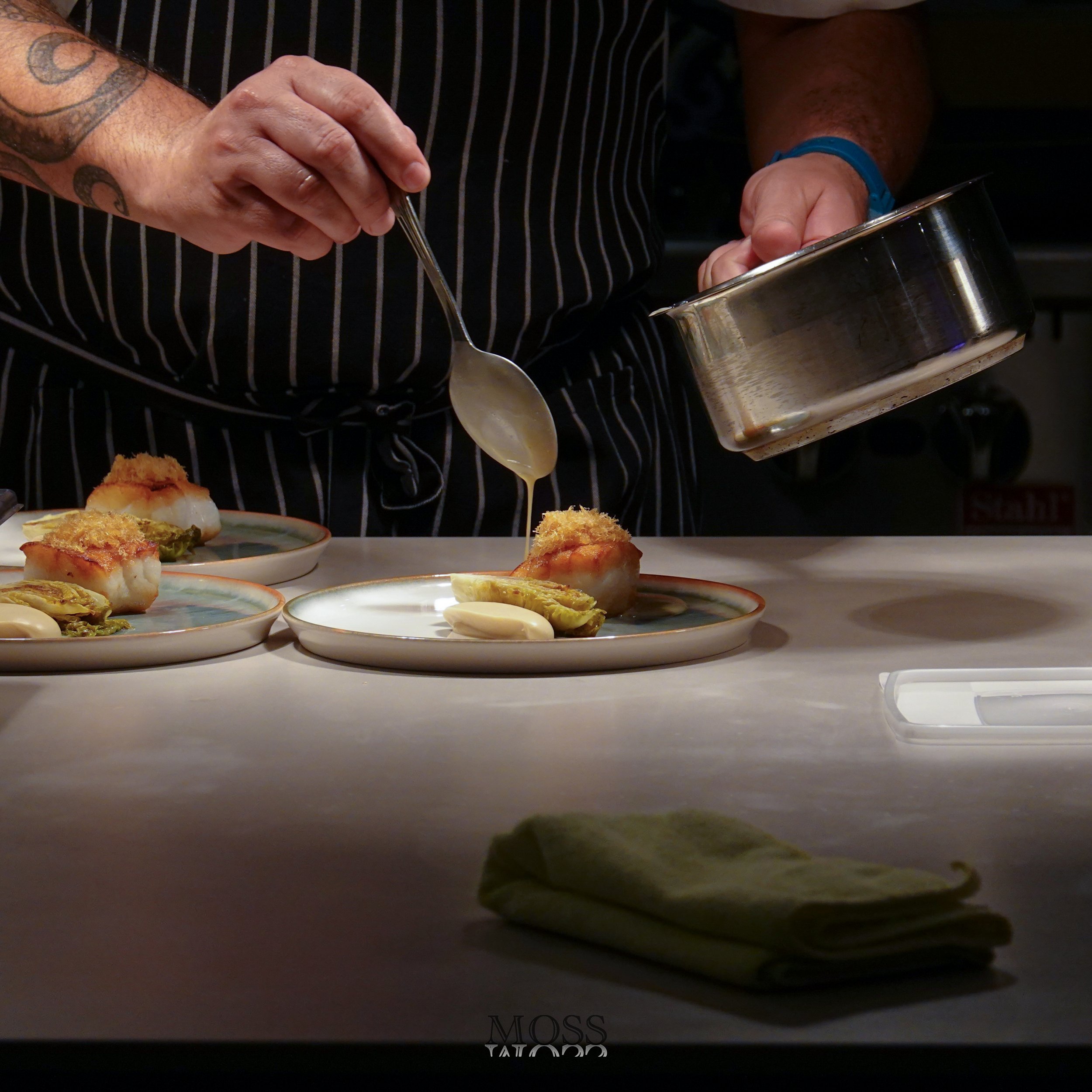 A chef in a striped apron drizzles sauce onto plated food, with several prepared plates with food, a green towel, and a container on the counter.
