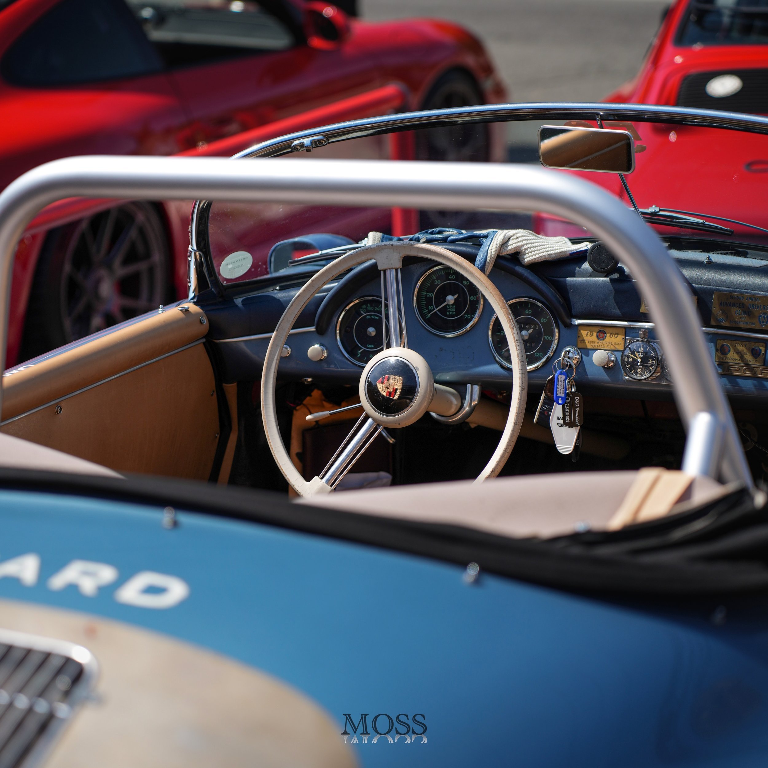 Interior of a vintage Porsche convertible sports car with a tan dashboard, wooden steering wheel, and classic gauges, parked next to a red sports car.
