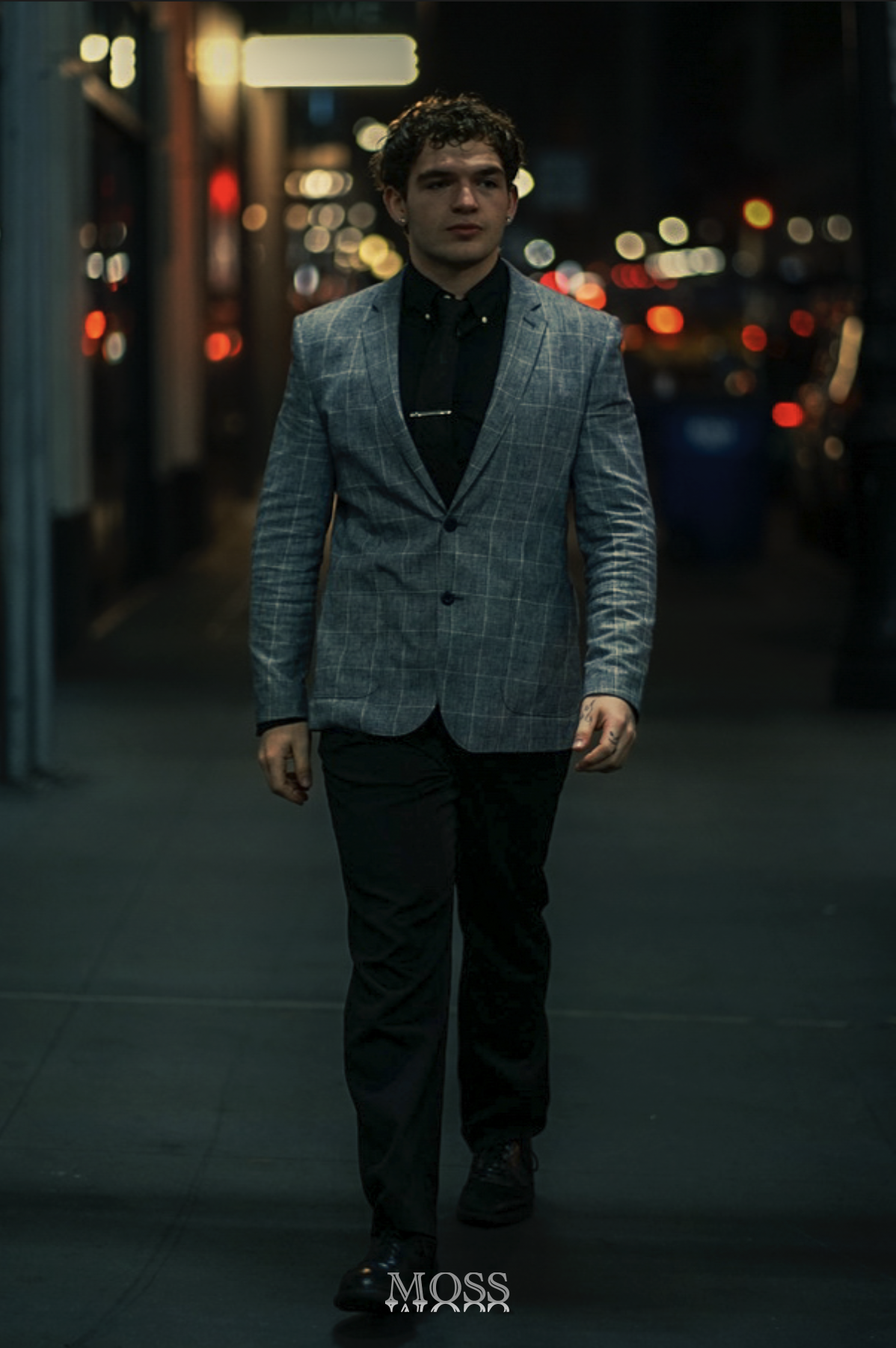 A man walking on a city street at night, wearing a gray checkered blazer, black shirt, black pants, and black shoes, with blurred city lights in the background.
