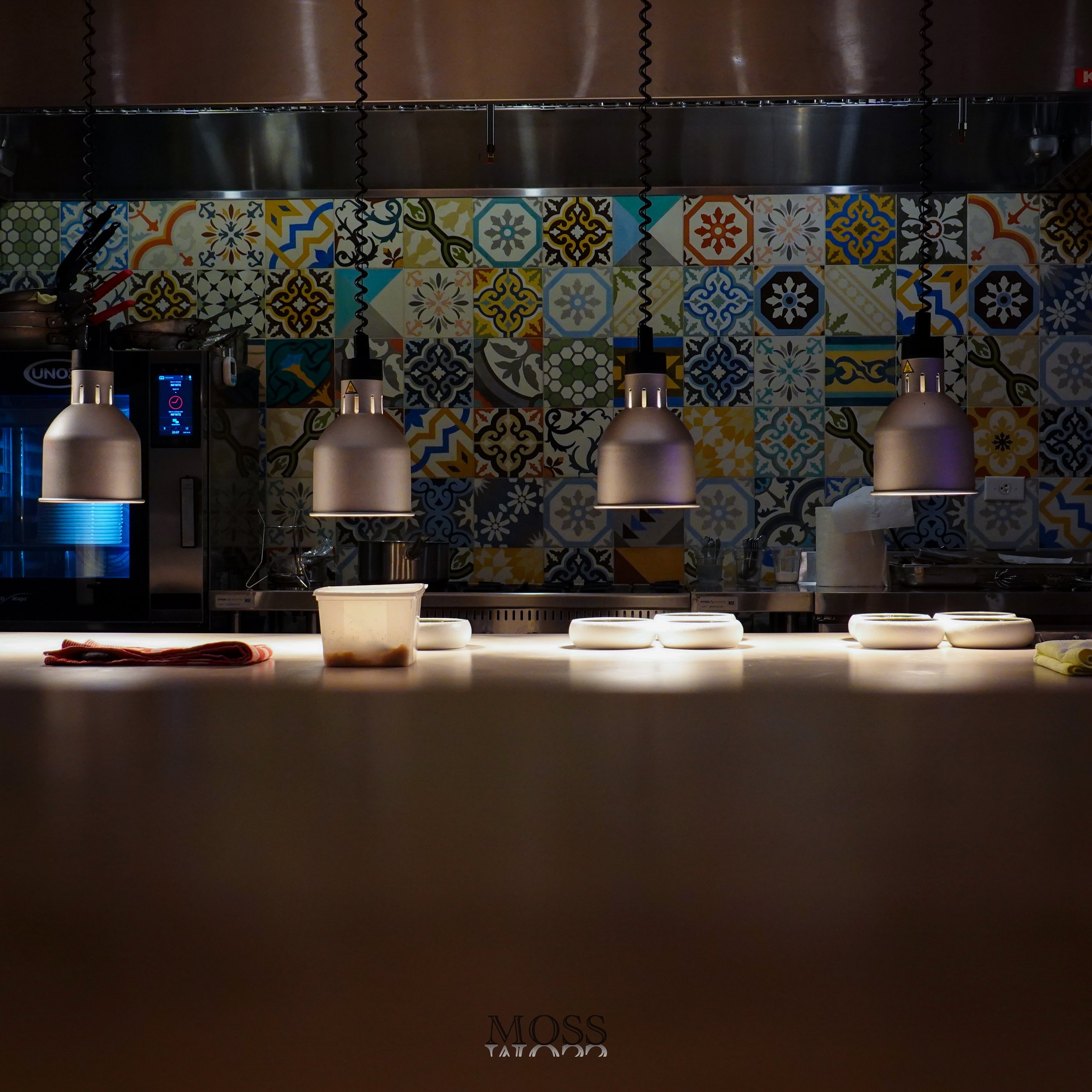 A kitchen with decorative colorful tile backsplash, four hanging pendant lights, and a countertop with some bowls and a container. The background includes a microwave oven and various kitchen utensils.