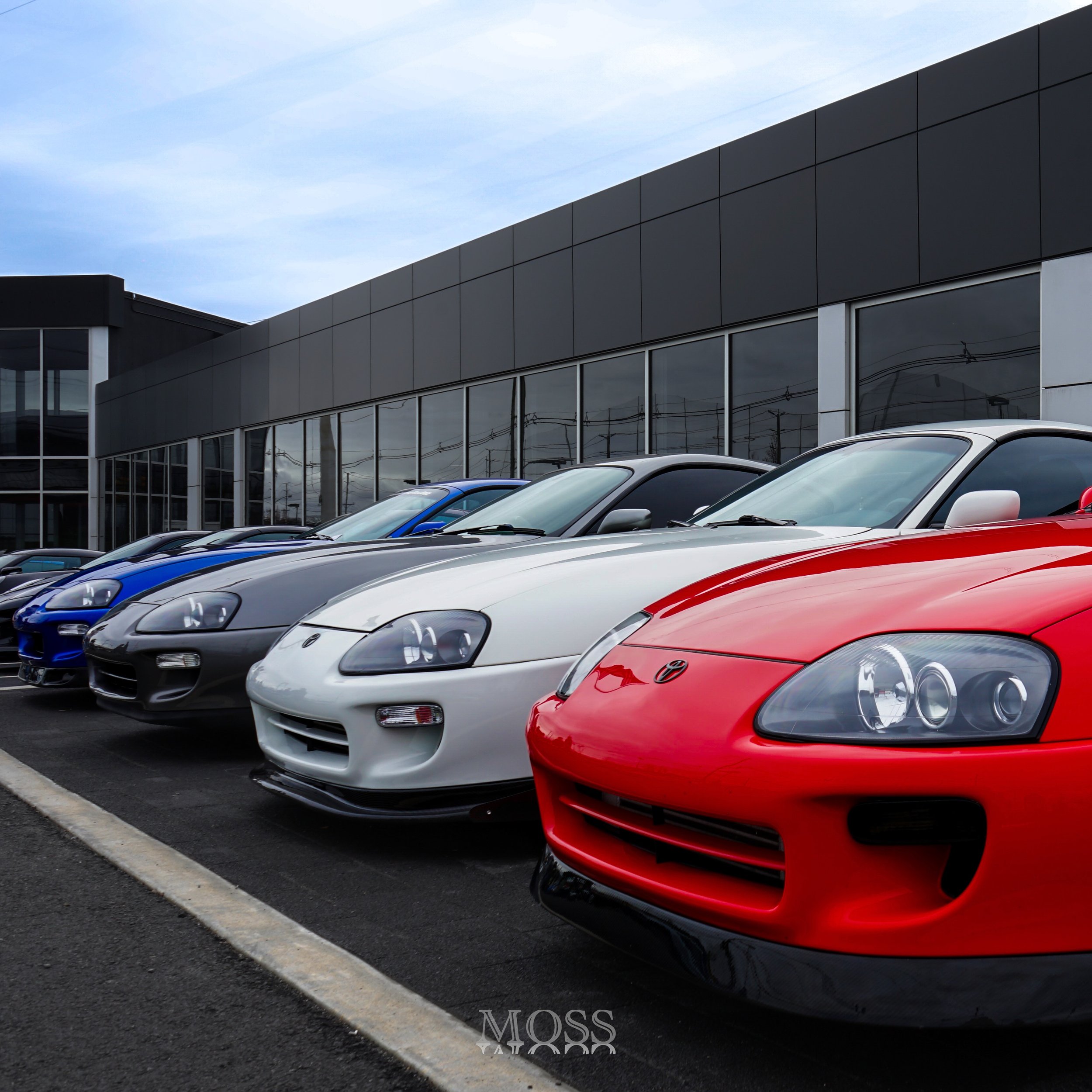 Line of four sports cars parked outside a modern glass building, with a blue sky above.