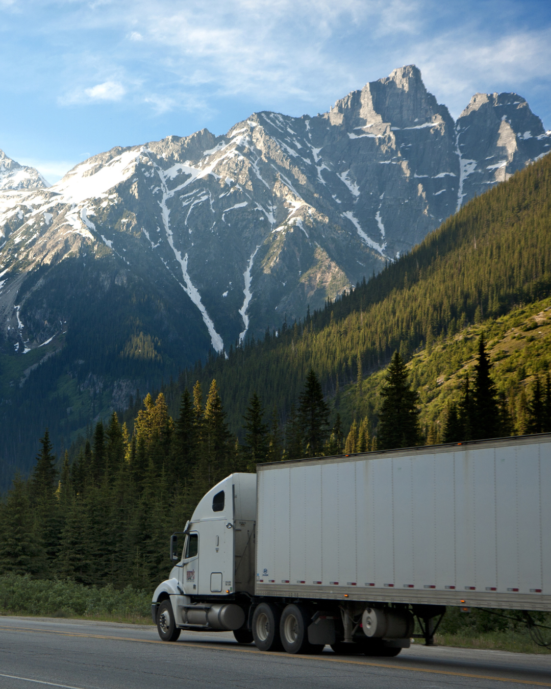 A white semi-truck driving on a highway with a backdrop of tall, snow-capped mountain peaks and a forest of pine trees.