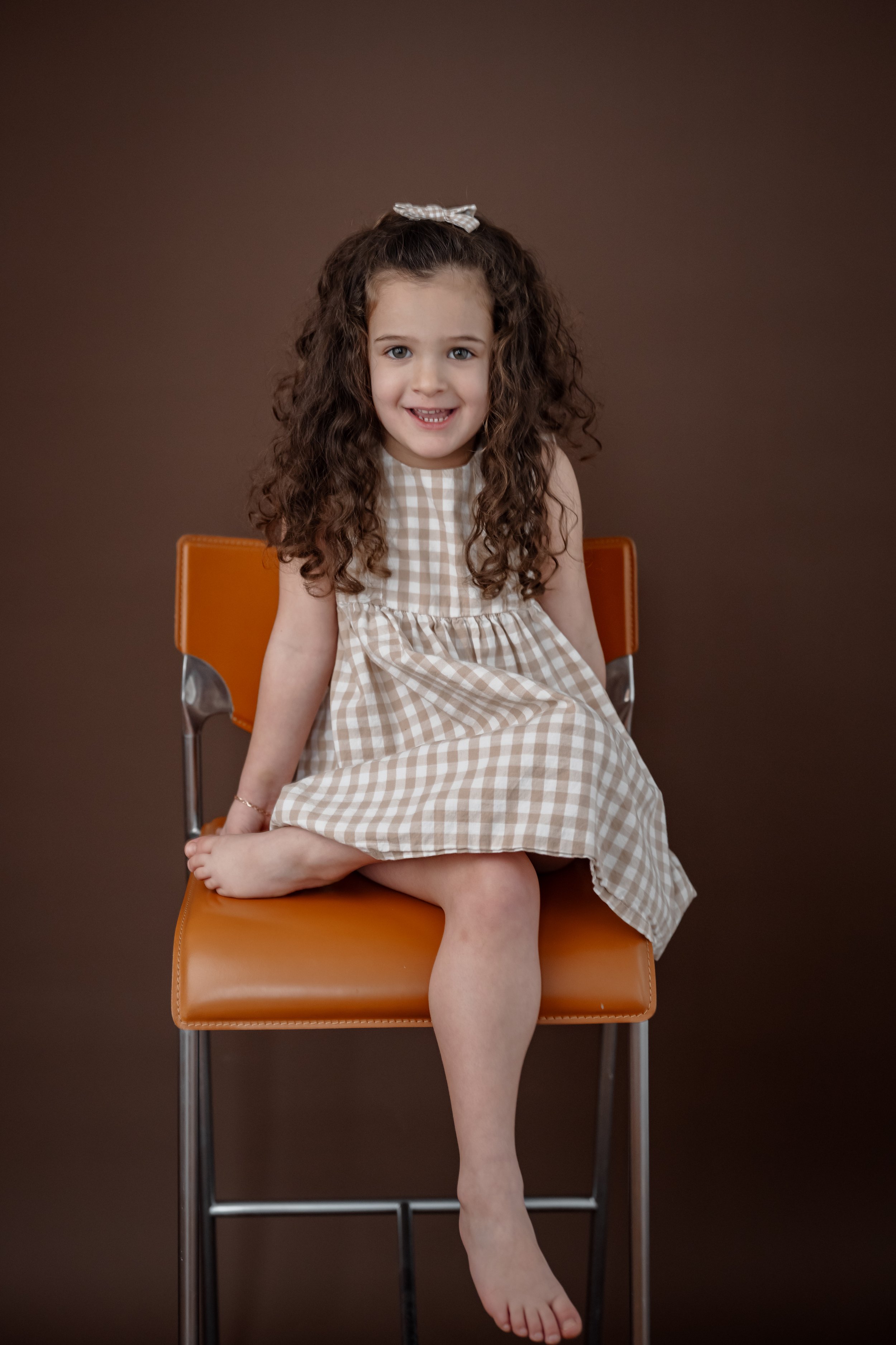 A young girl with curly brown hair, wearing a beige and white checkered dress and a matching bow, sitting on an orange chair against a brown background.