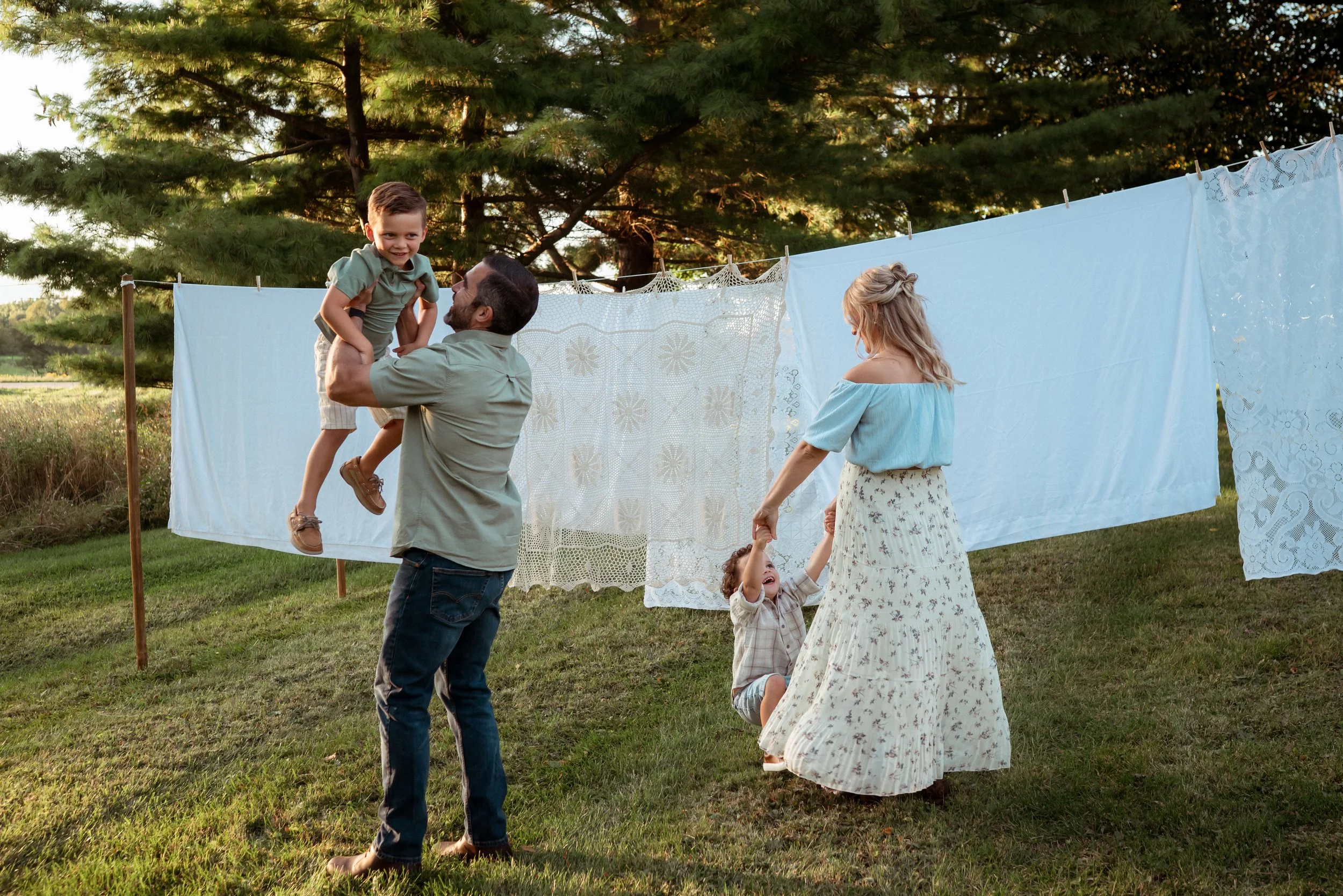 A family playing together outdoors during a summer photoshoot, with a father lifting his young son and a mother spinning with their toddler in front of hanging linens and lace. Soft evening light, greenery, and vintage textures create a warm, nostalg