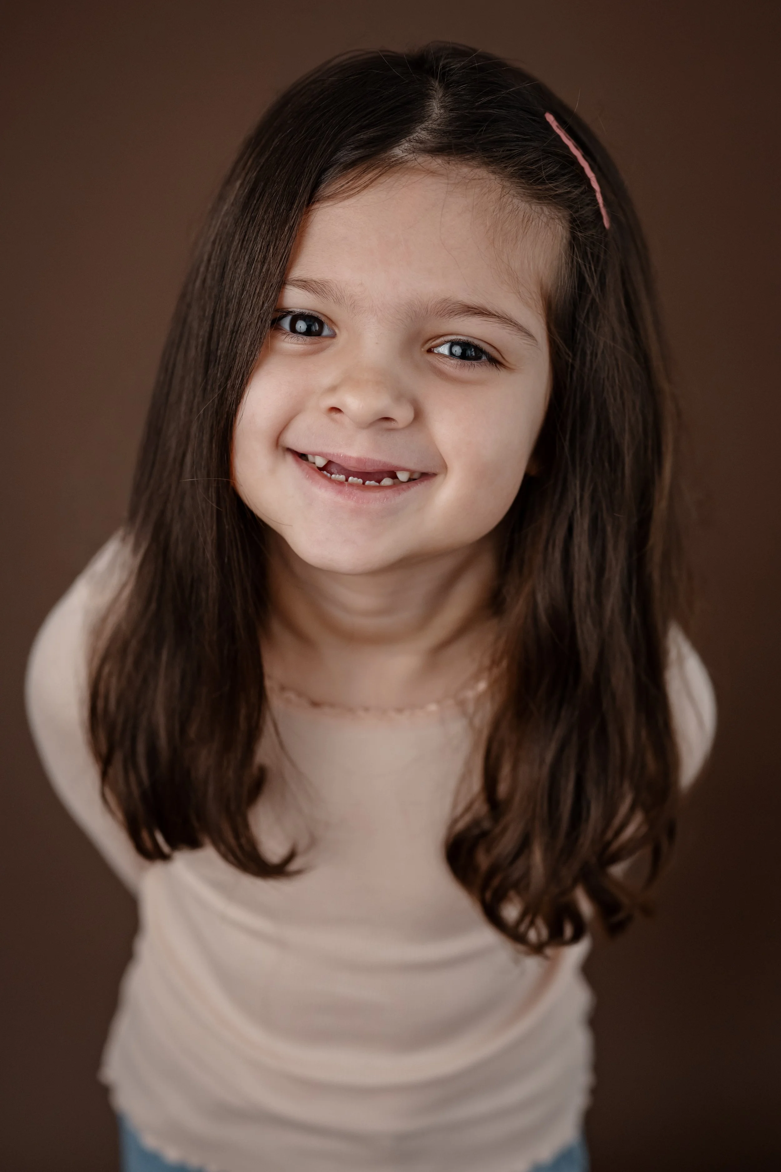 Young girl with long dark hair smiles at the camera during a child portrait session against a warm brown studio backdrop.