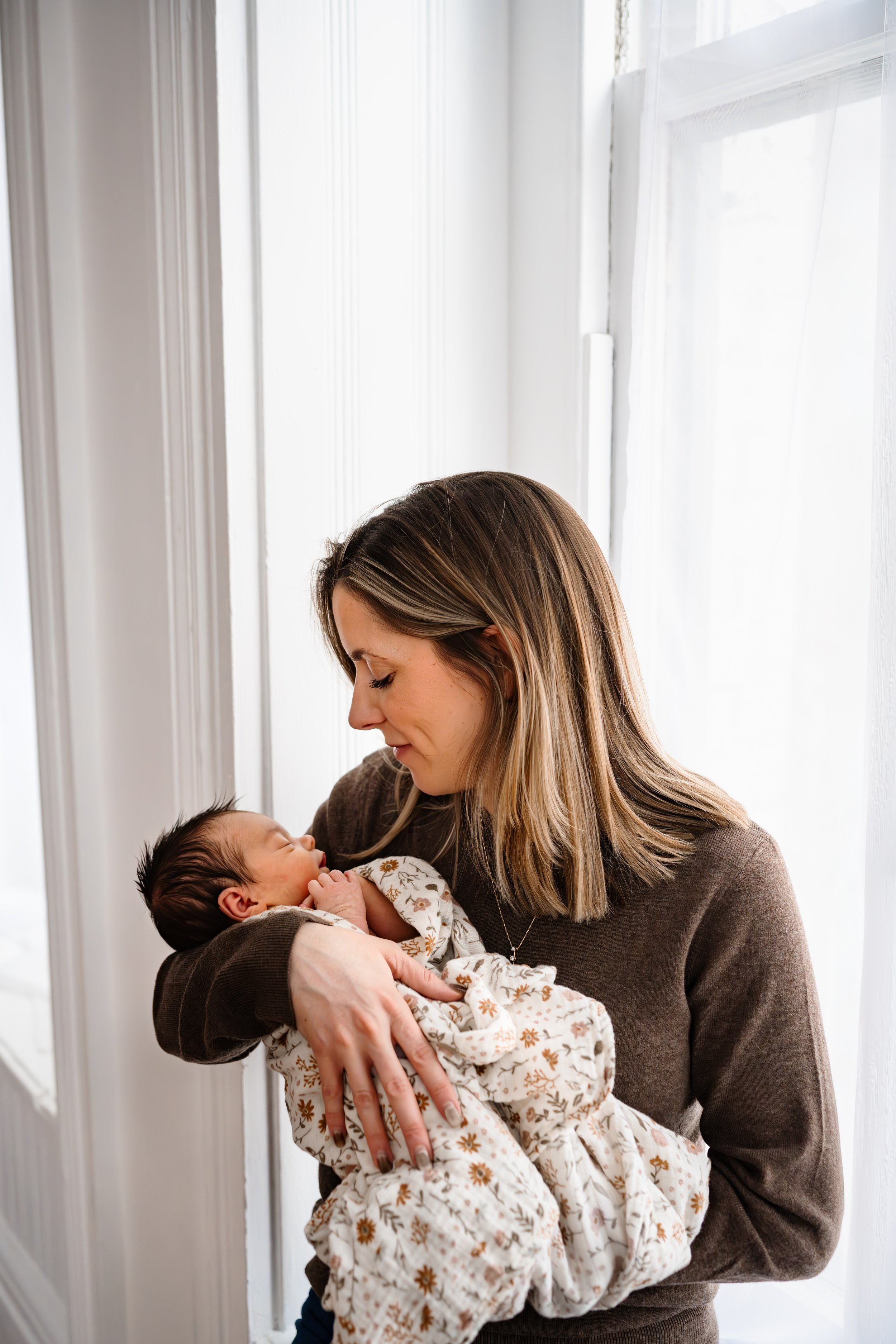 Mother cradles her sleeping newborn in her arms beside a bright window, gazing down with a gentle smile while the baby is wrapped in a soft floral swaddle.