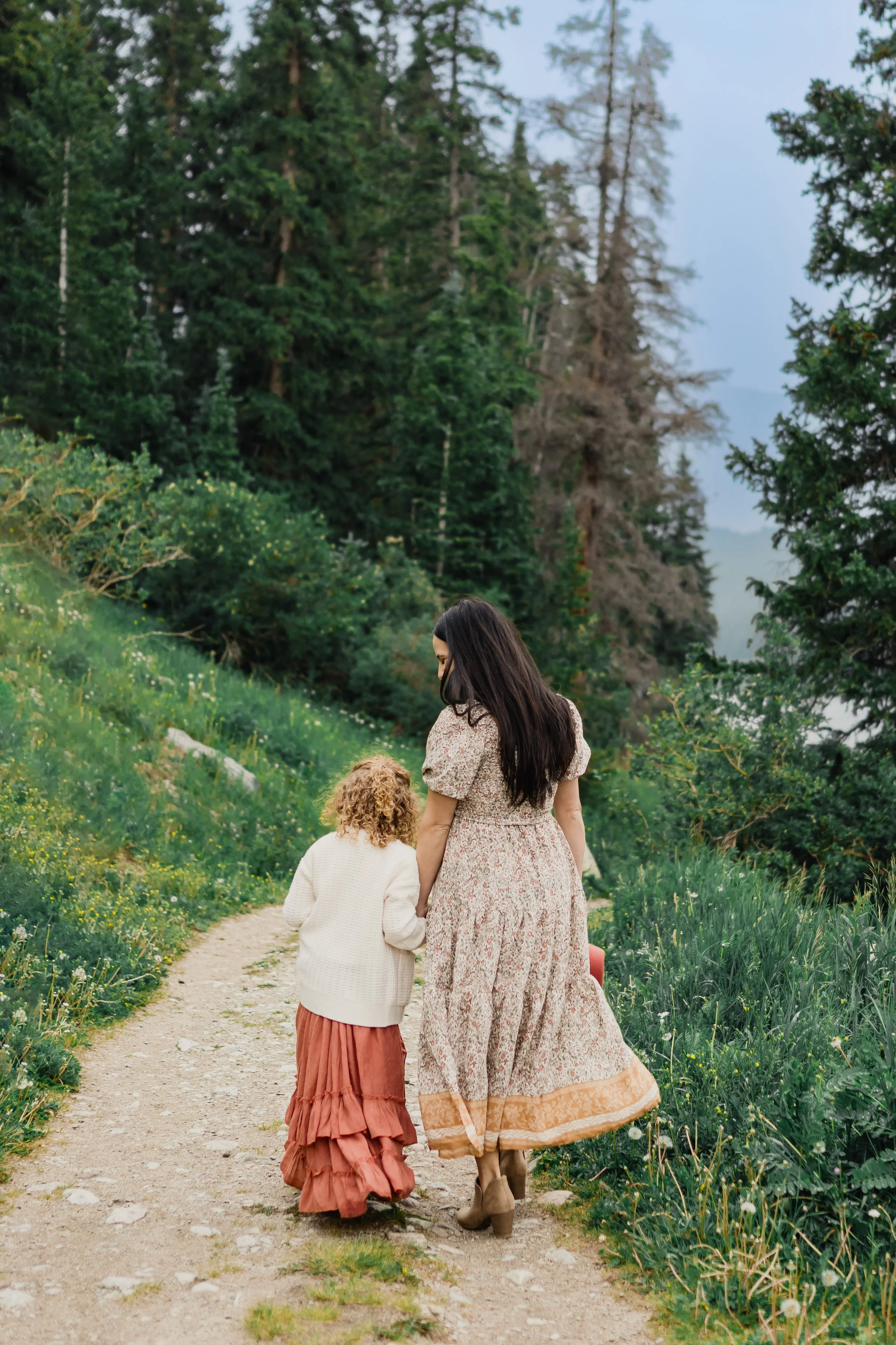 A mother and her young child walk hand in hand along a winding forest trail surrounded by lush greenery and tall pine trees. Seen from behind, they wear flowing neutral-toned clothing, creating a peaceful, storytelling moment in nature.
