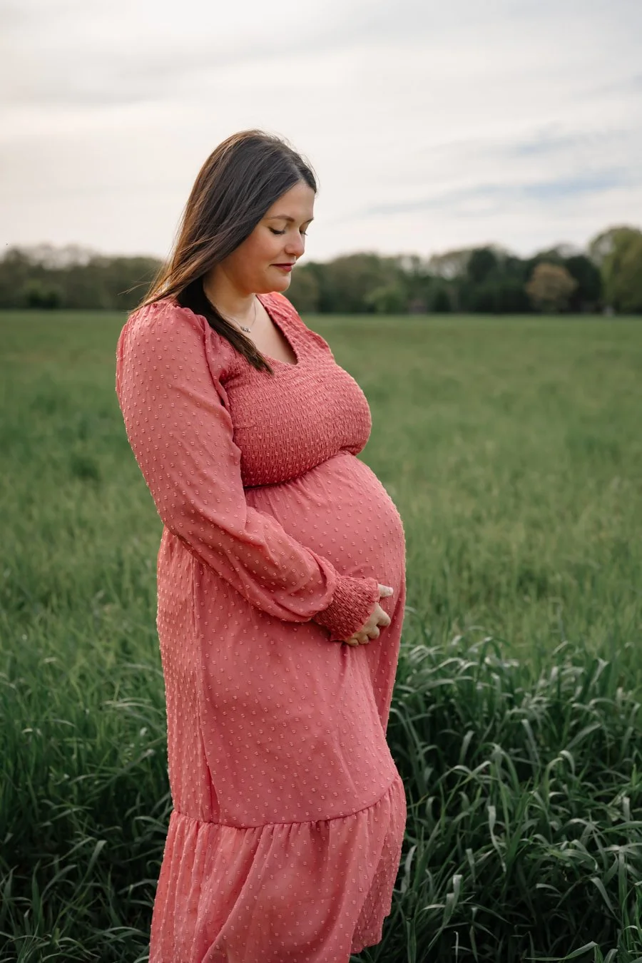 Expectant mother stands in a lush green field wearing a flowing coral dress, gently cradling her baby bump while looking down, creating a peaceful outdoor maternity portrait under a soft evening sky.