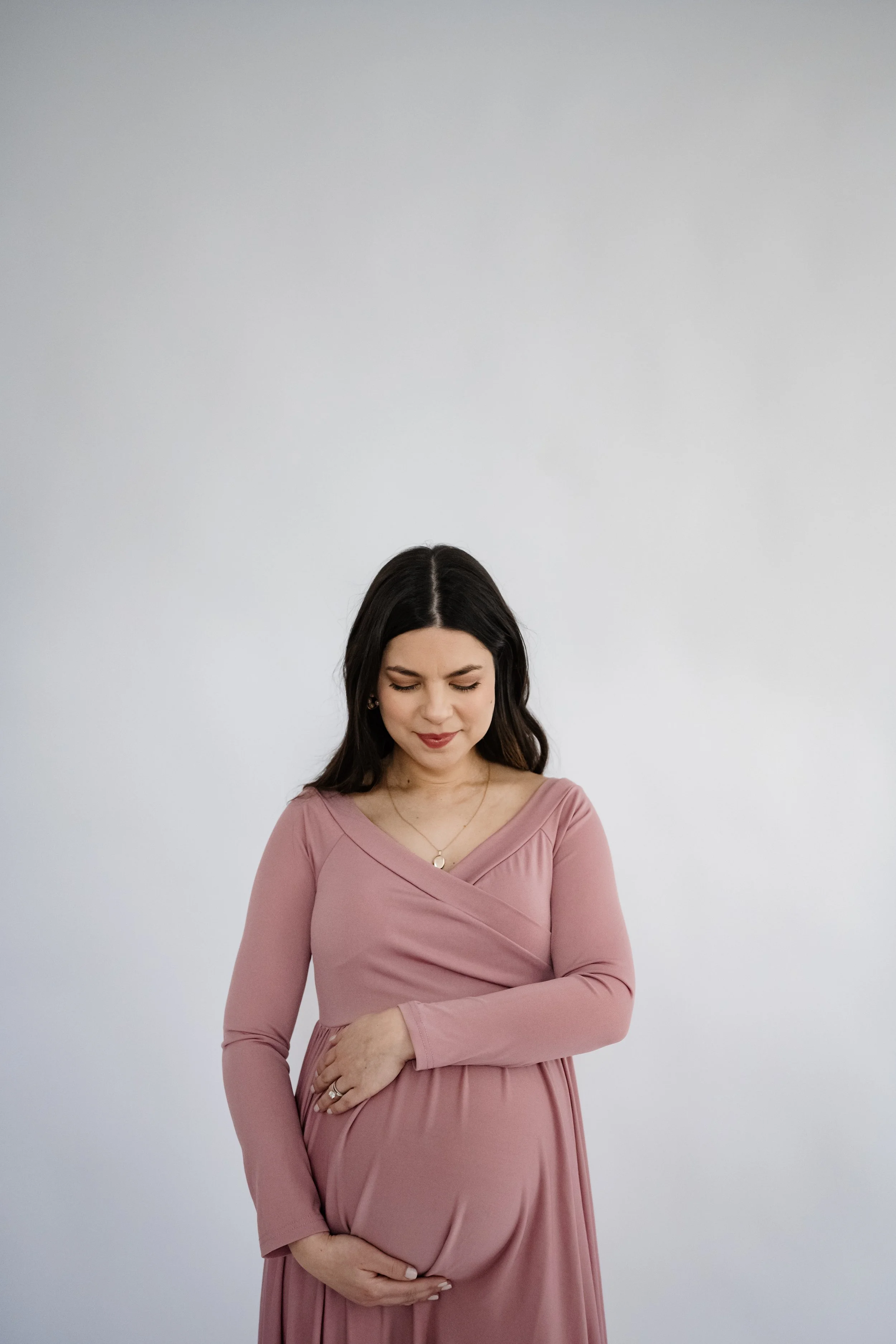 A pregnant woman in a dusty rose maternity dress stands against a bright white studio backdrop, gently holding her baby bump with both hands. Looking down with a peaceful expression, the minimalist portrait highlights the beauty and anticipation of p