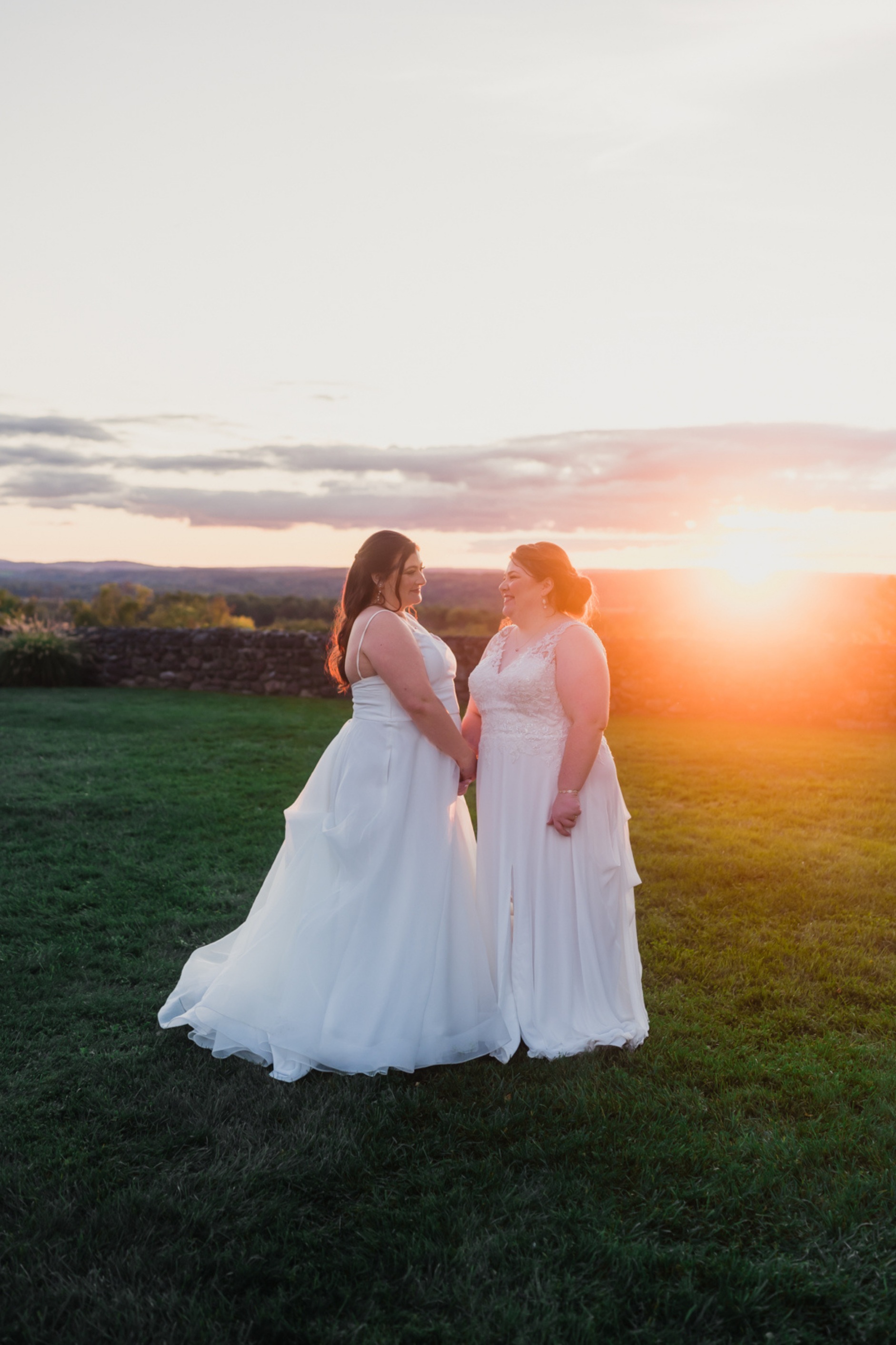 Two brides standing hand in hand outdoors at sunset during a wedding portrait, wearing white gowns and smiling at each other as warm light fills the scene.