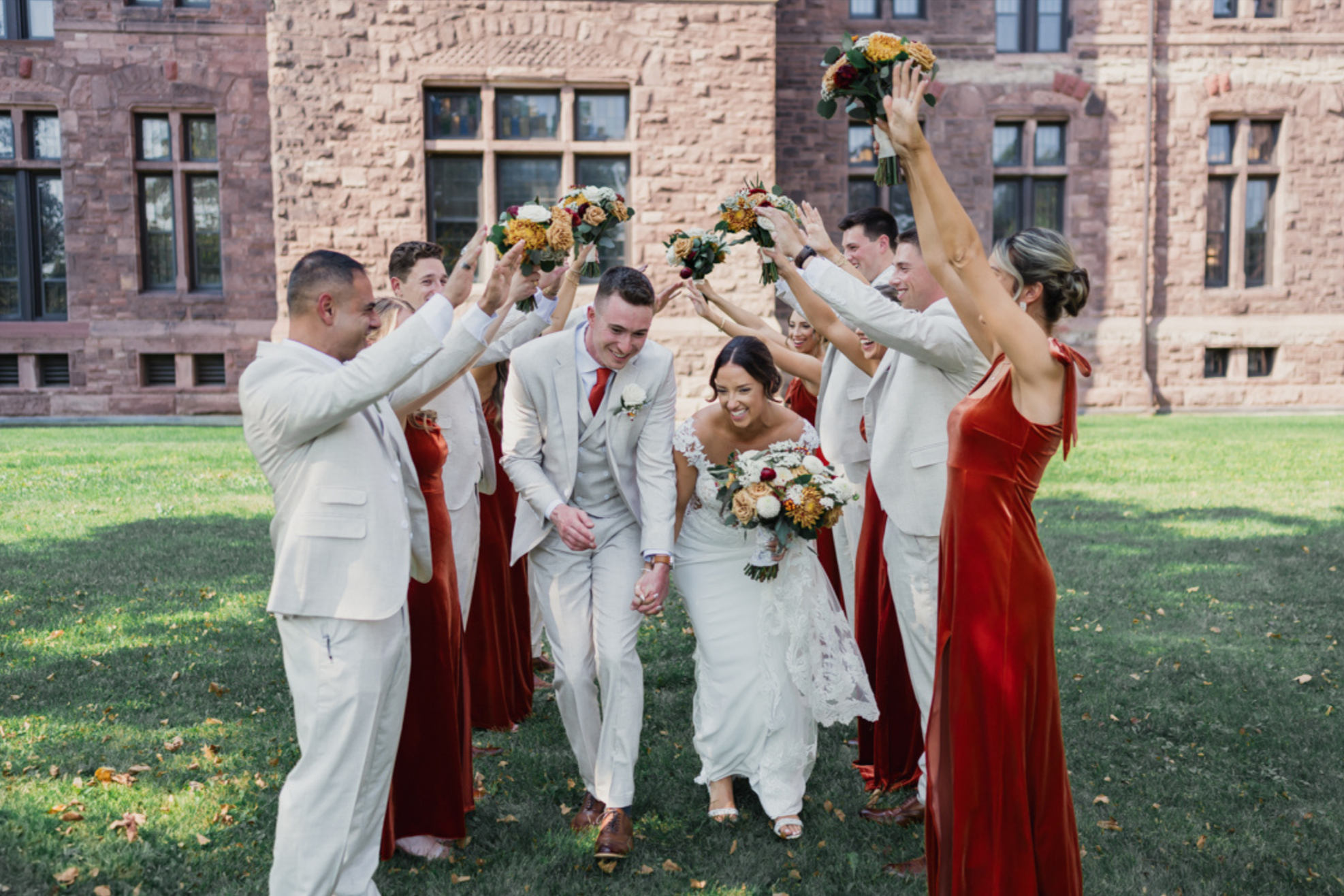 A bride and groom walking hand in hand beneath raised bouquets held by their wedding party, smiling and laughing during an outdoor wedding celebration in front of a historic stone building.
