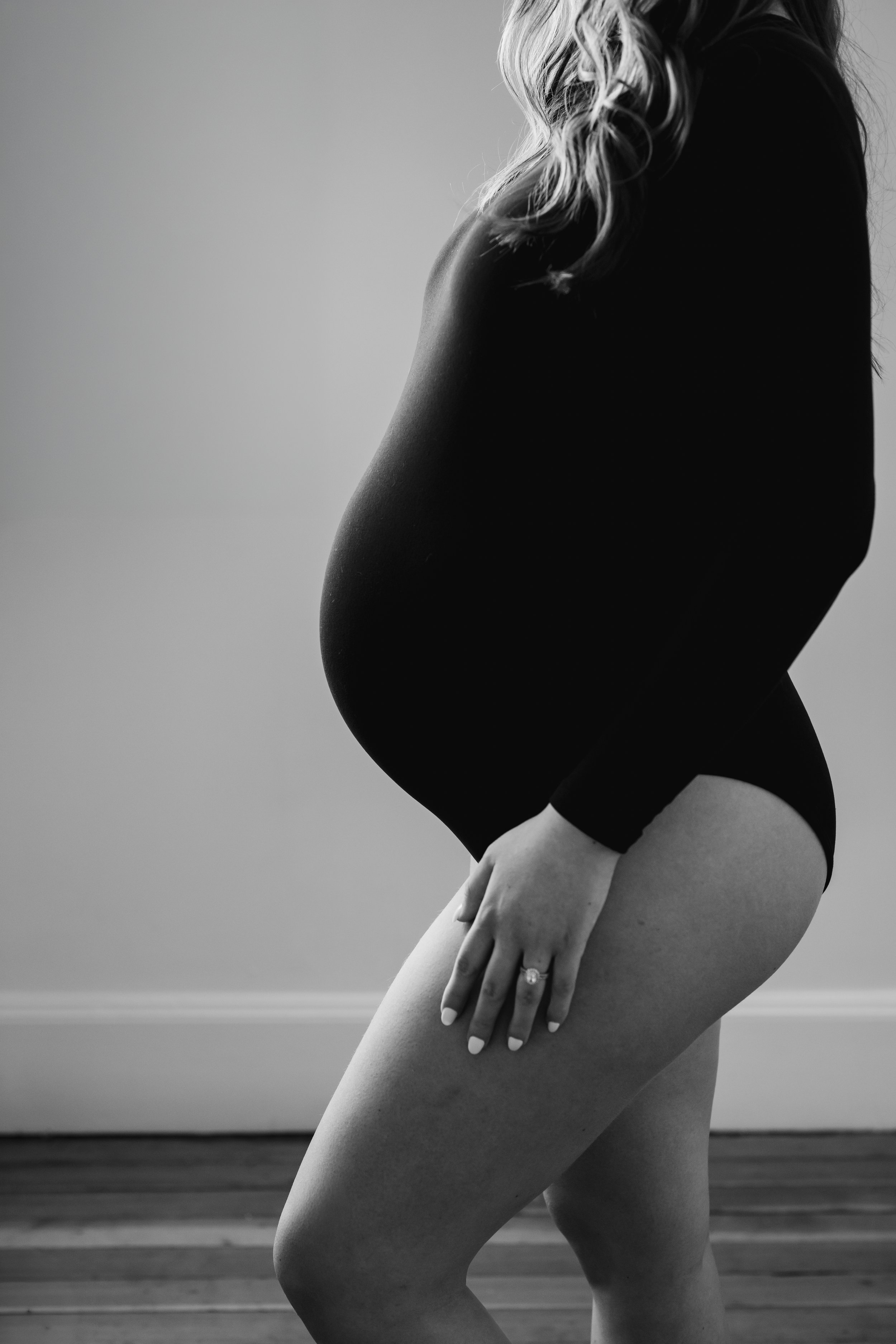 Black and white side profile of a pregnant woman in a fitted bodysuit, highlighting her baby bump in a simple, softly lit indoor setting.
