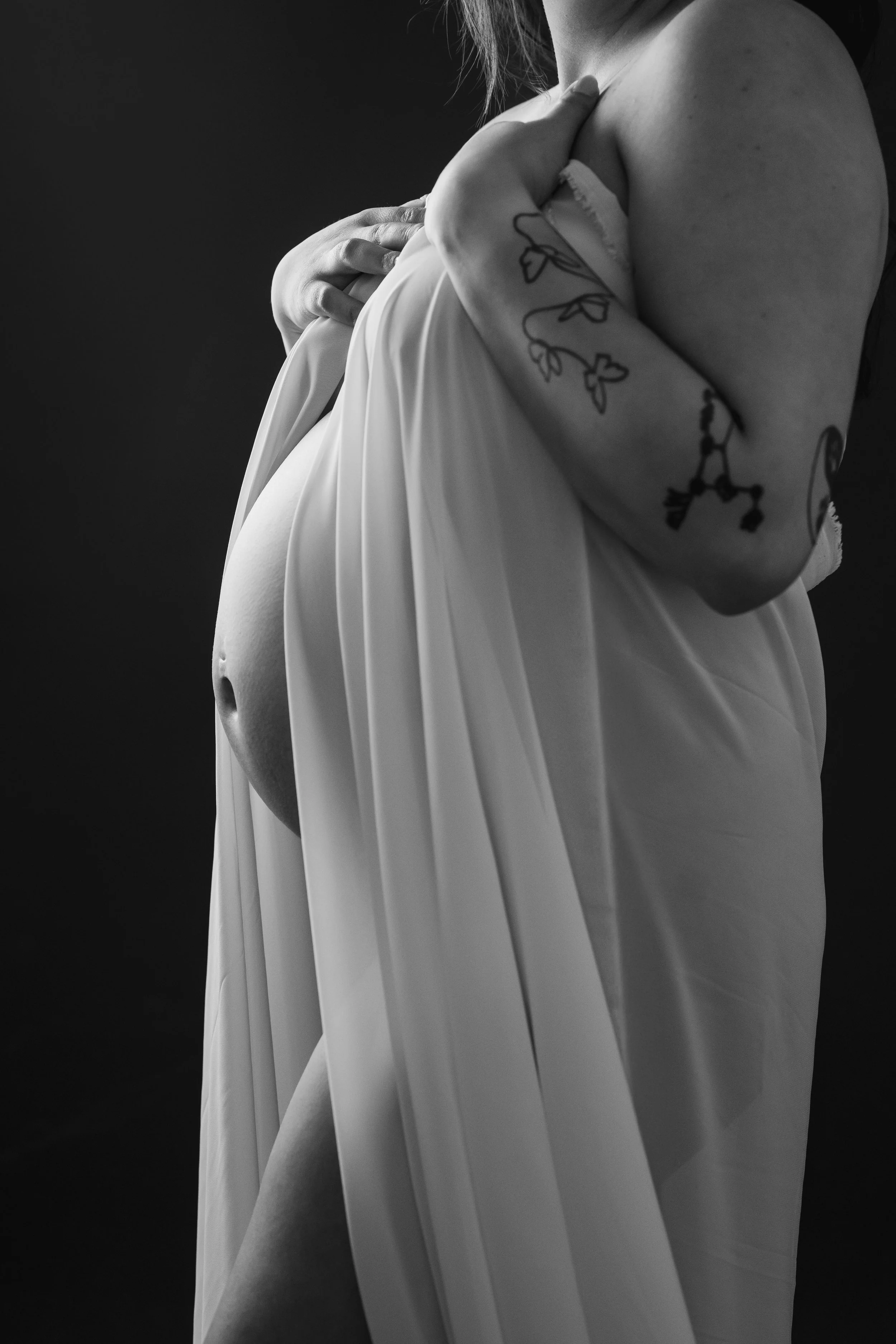A black and white photograph of a pregnant woman, focusing on her upper body and belly, holding a fabric near her chest with tattoos on her arm visible.