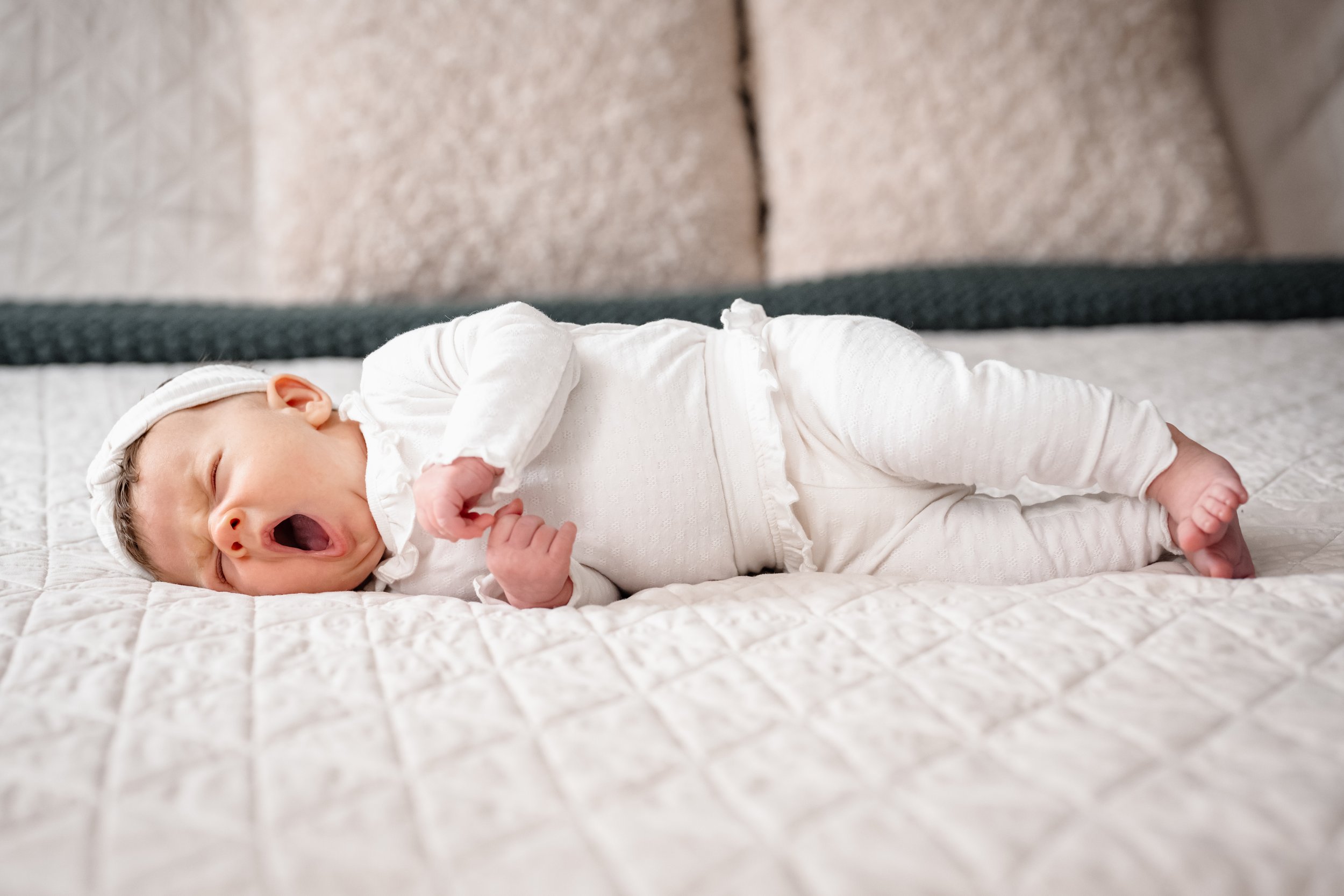 A newborn baby lying on a bed in a white textured outfit and headband, captured mid-yawn during an in-home newborn photography session with soft, neutral tones.