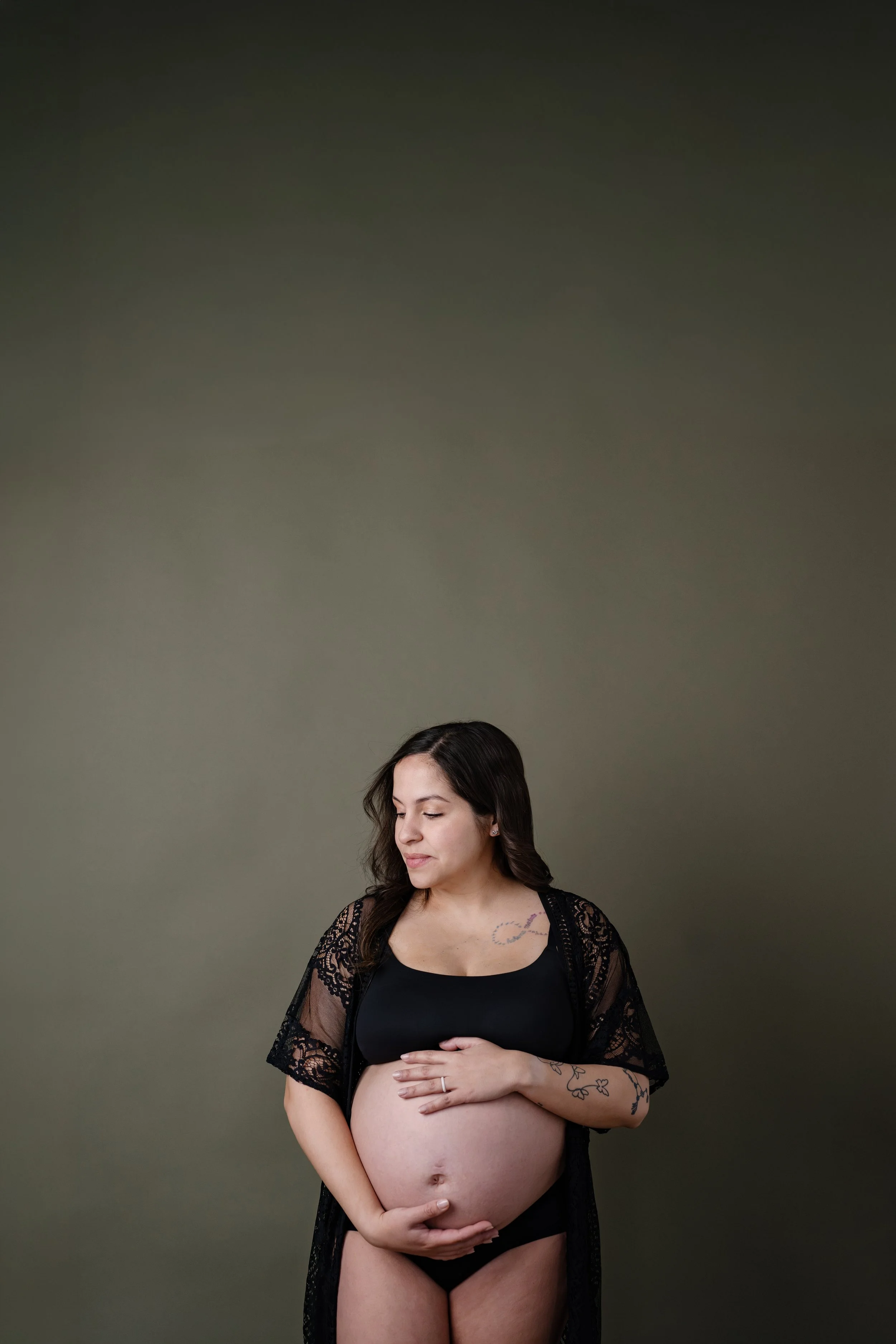 Pregnant woman standing against a muted green backdrop, gently holding her baby bump while wearing a black lace robe and looking down with a calm expression.