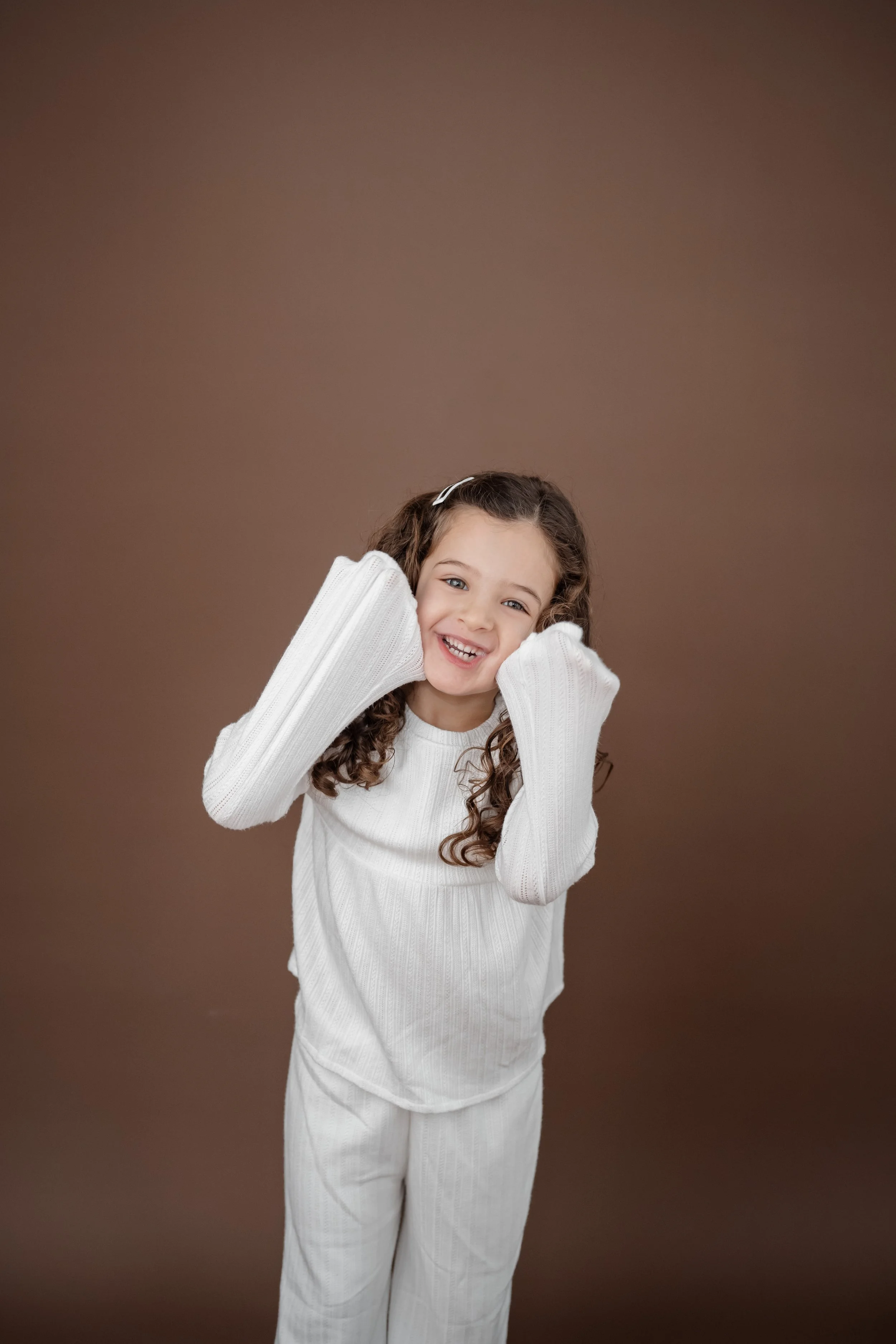 A young girl with curly brown hair, smiling and wearing a white sweater and pants, standing against a brown background.